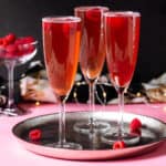 Pink Champagne cocktail with raspberries in flute glasses on a silver platter and pink and black background.