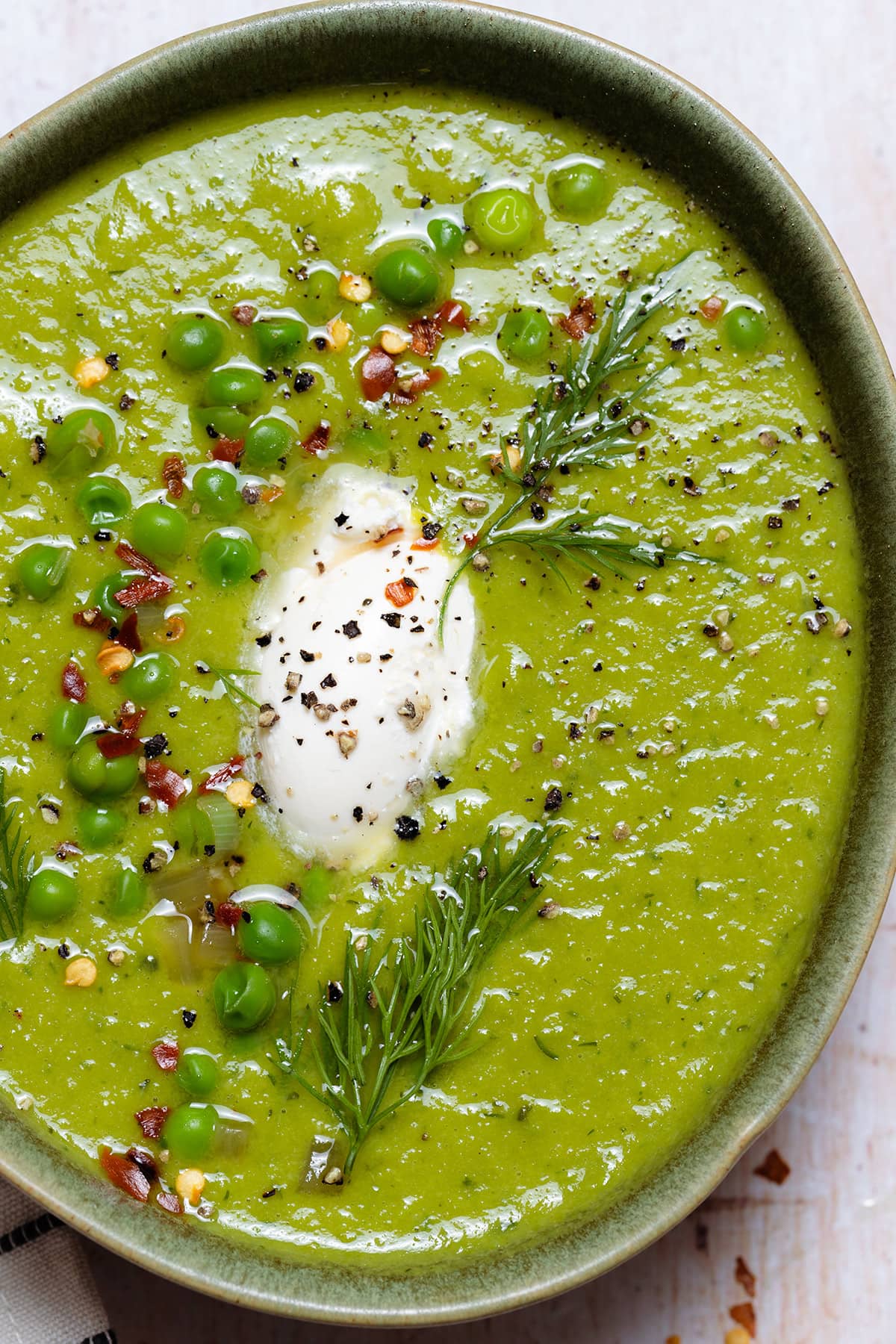 A close up of green bowl with green soup garnished with peas, fresh, dill, chili flakes, and creme fraiche.