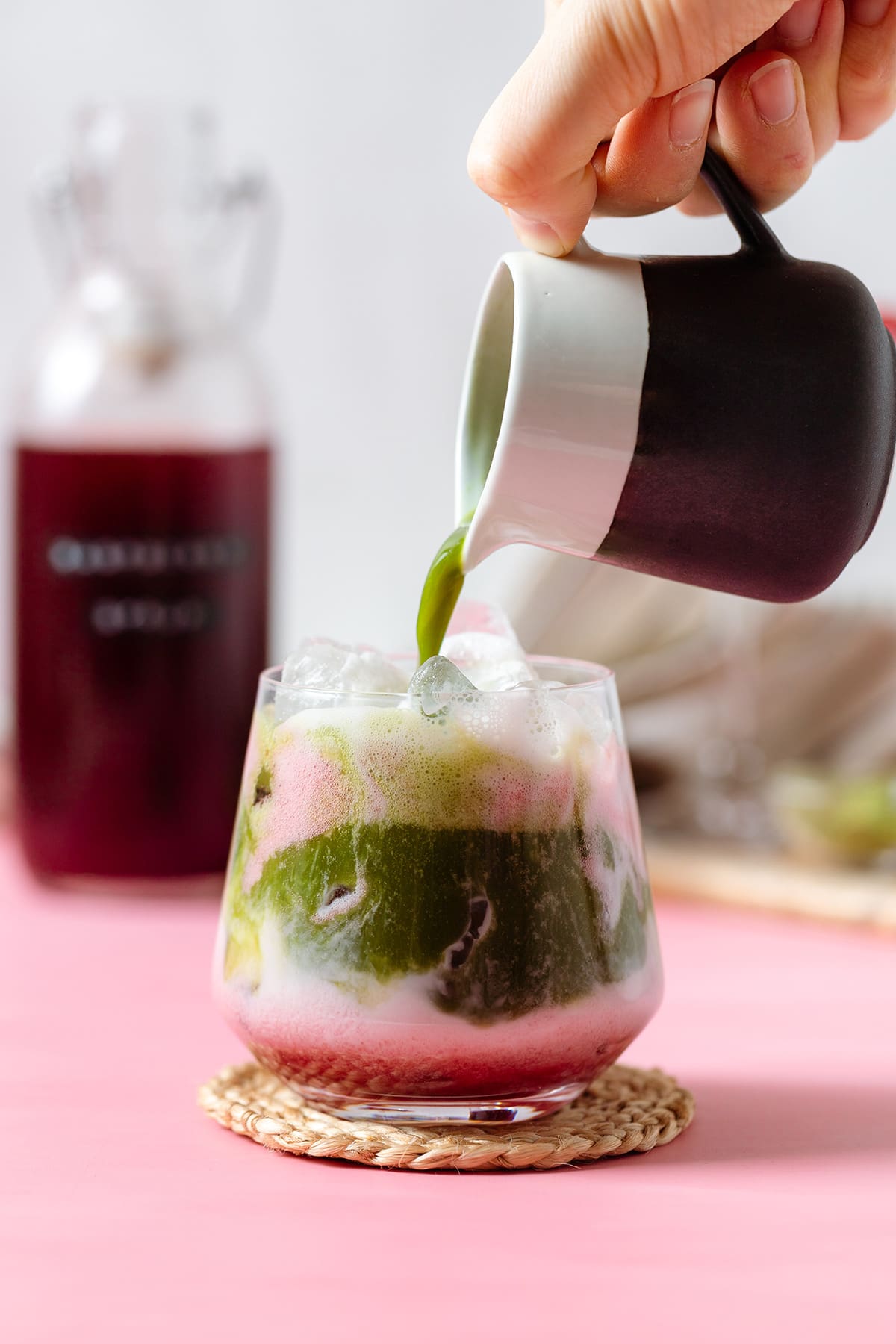 Matcha being poured into a glass with almond milk and raspberry syrup on a pink background.