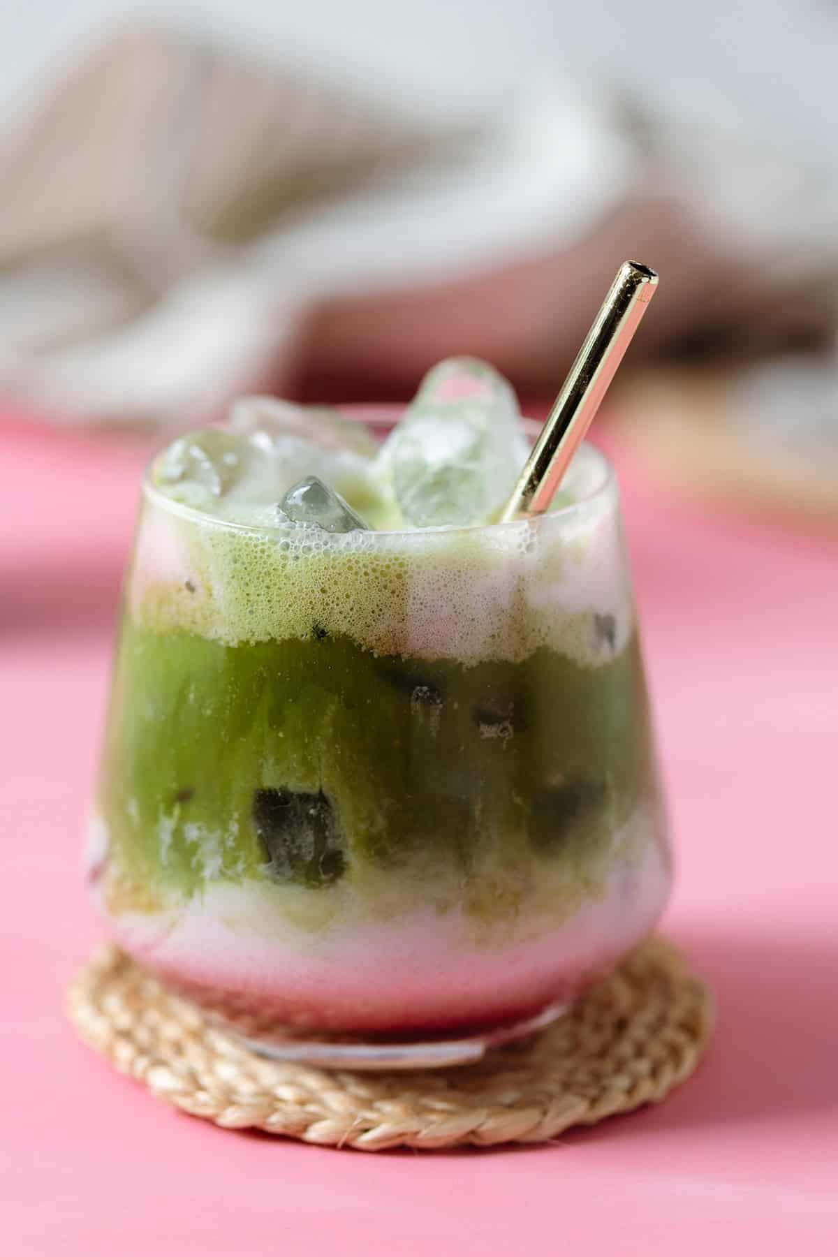 A glass with matcha, raspberry syrup, almond milk, and a straw on a pink background.