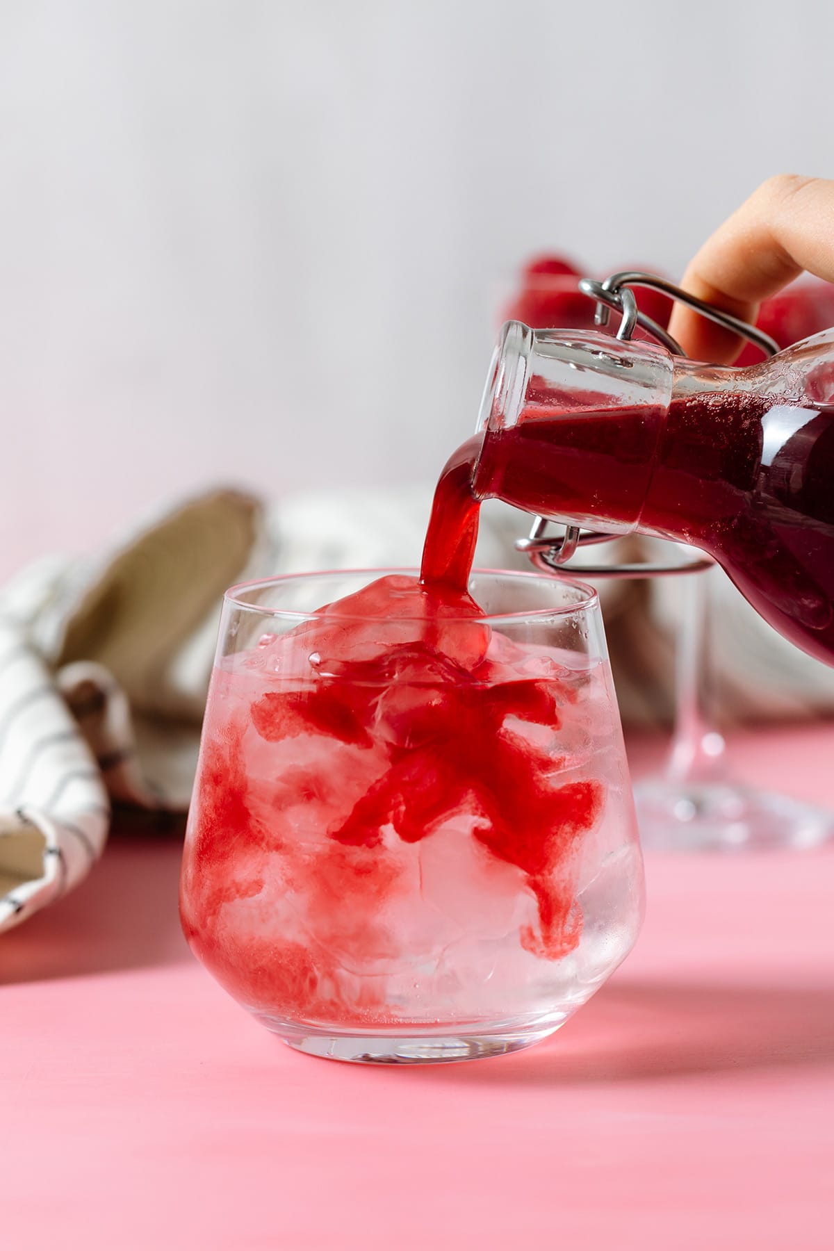 Raspberry syrup being poured into a glass of water on a pink background.