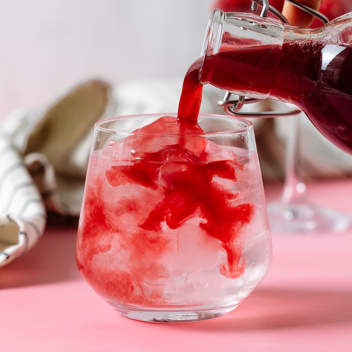 Raspberry syrup being poured into a glass of water on a pink background.