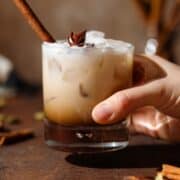A hand holding a White Russian cocktail in a double old fashioned glass garnished with a cinnamon stick and star anise on a wooden backdrop.