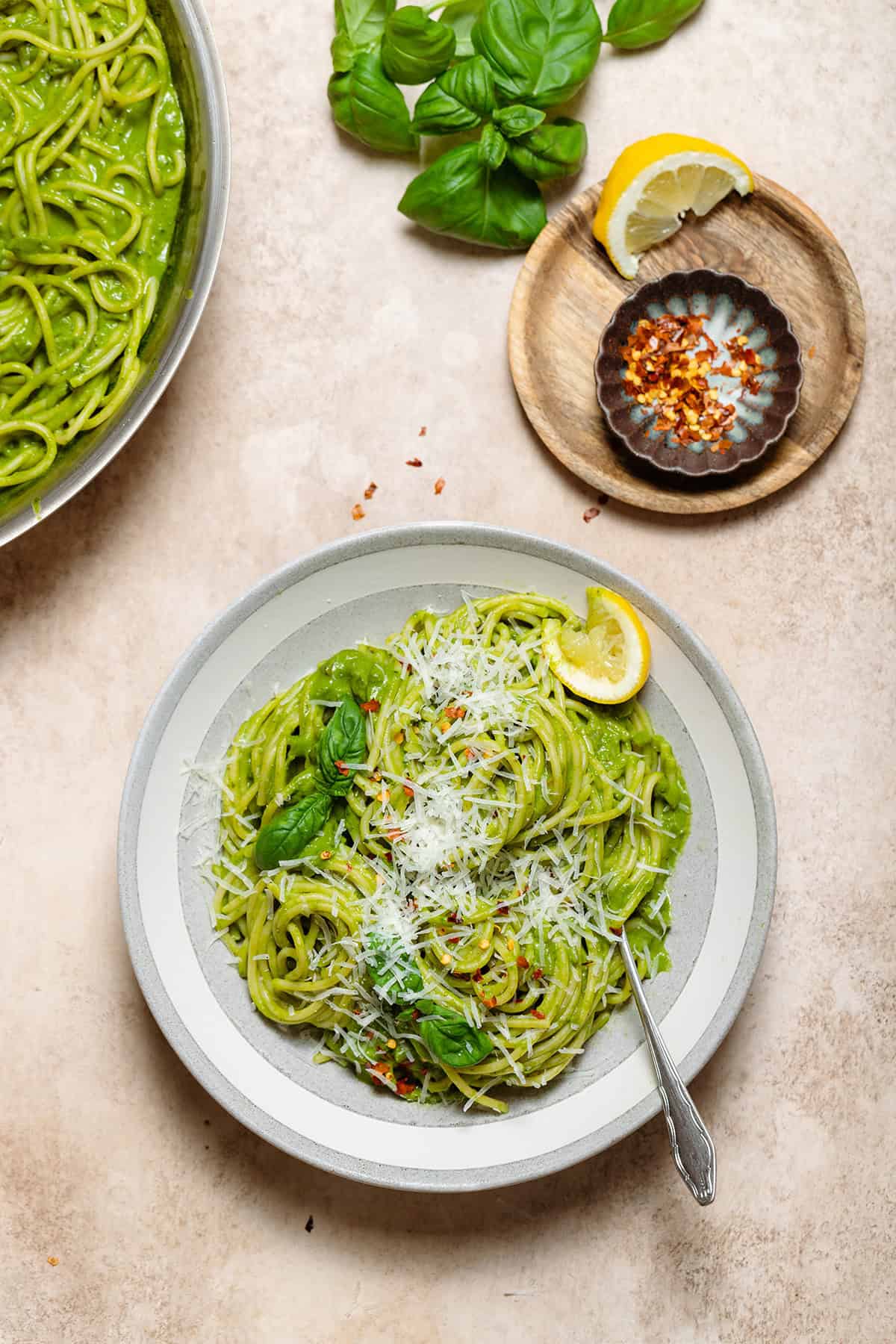 Spaghetti with green sauce in a low grey bowl garnished with fresh shredded parmesan, basil, chili flakes, and lemon.