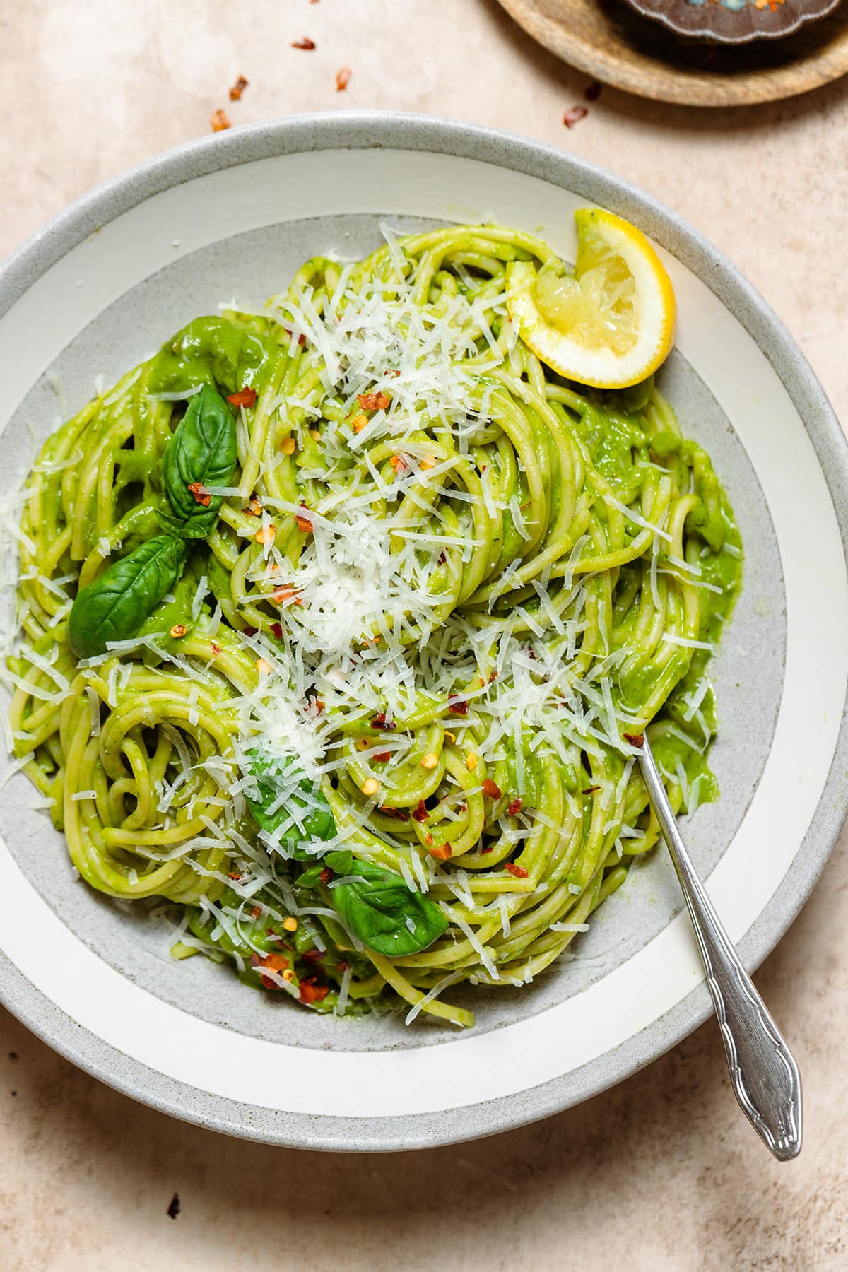 Spaghetti with green sauce in a low grey bowl garnished with fresh shredded parmesan, basil, chili flakes, and lemon.