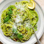 Spaghetti with green sauce in a low grey bowl garnished with shredded parmesan, fresh basil, chili flakes, and lemon.