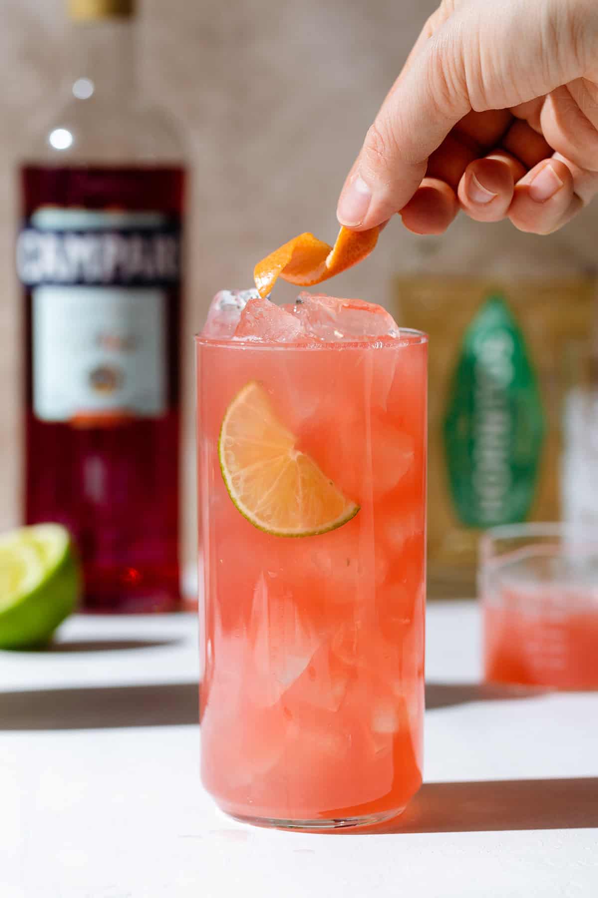 A hand placing an orange peel on a cocktail in a tall glass with ice.