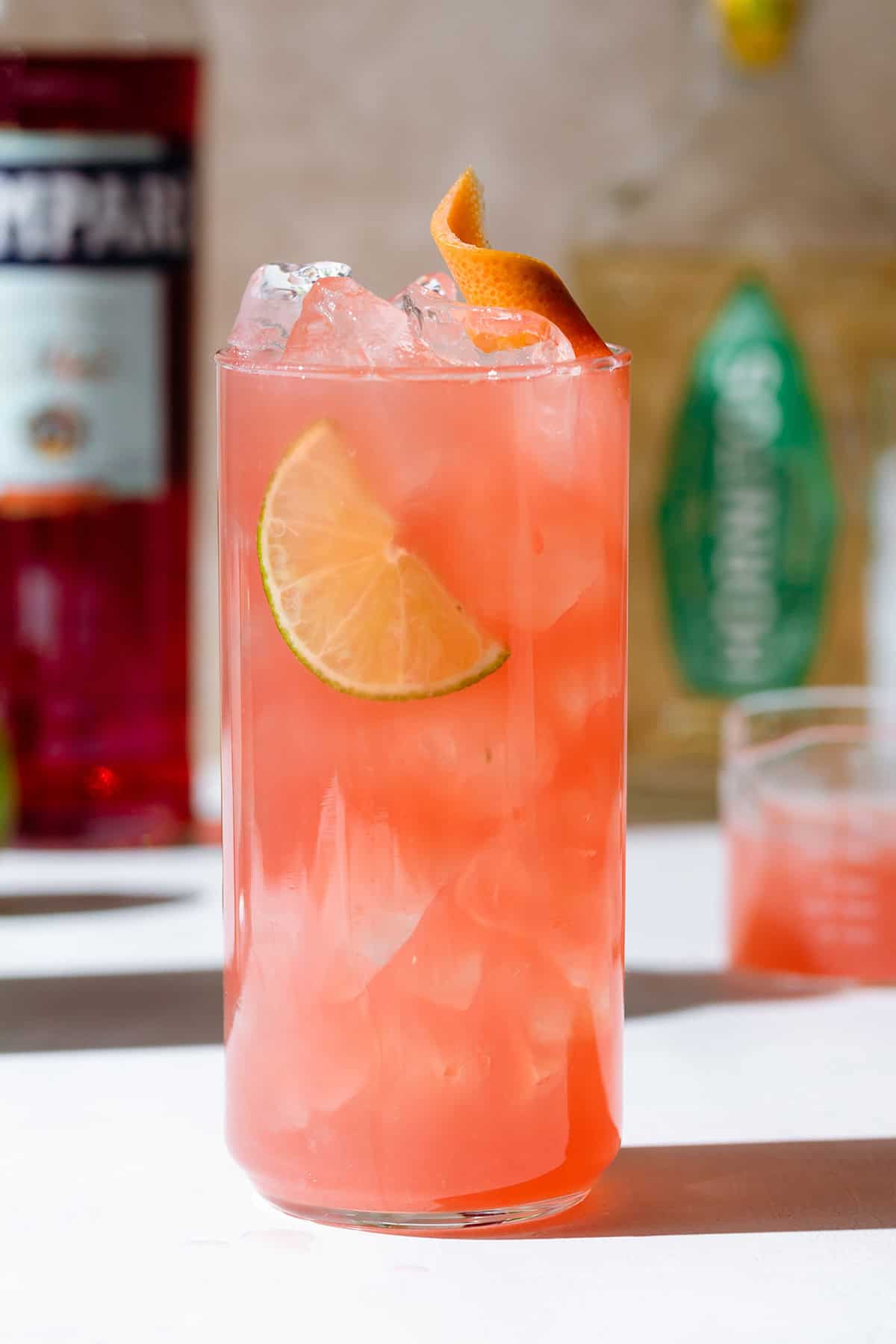 Light red colored campari cocktail in a tall glass with ice and a lime slice.