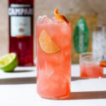 Peach colored campari cocktail in a tall glass with ice and a lime slice.
