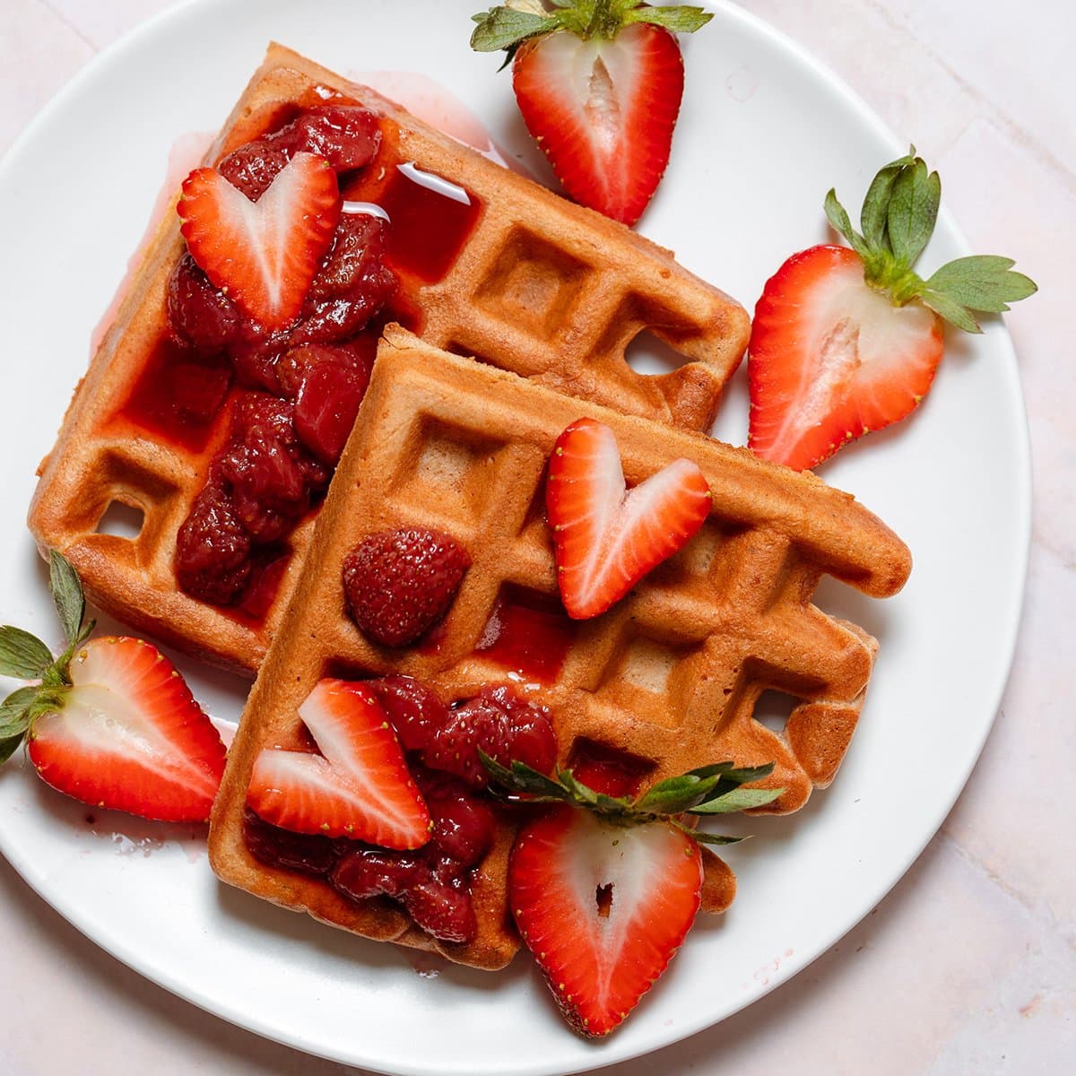 Waffles with stewed and fresh strawberries on a white plate on a stone background.