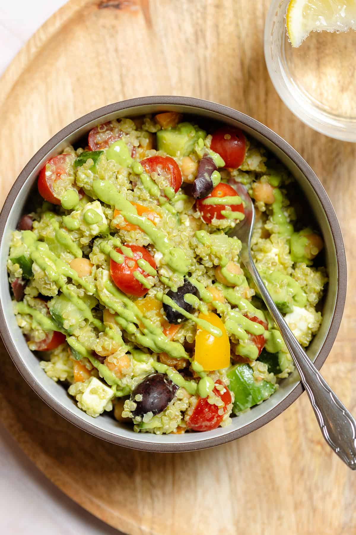 Colorful vegetable quinoa salad with green avocado dressing in a ceramic bowl on a wooden serving plate.