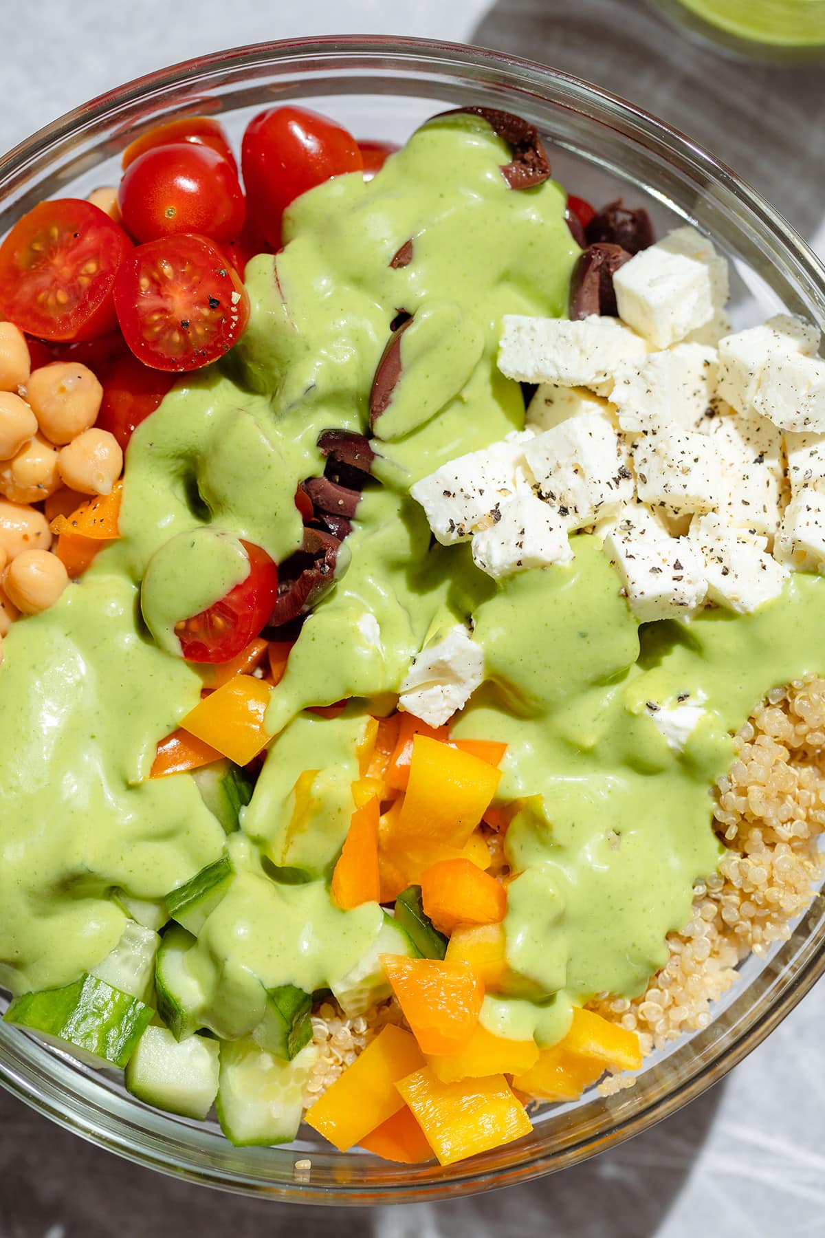 A close up of a glass bowl with quinoa, chickpeas, tomatoes, feta, olives, and green avocado dressing.