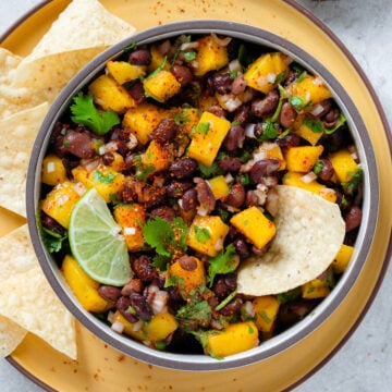 Black bean mango salsa in a bowl with tortilla chips around it on a yellow plate.
