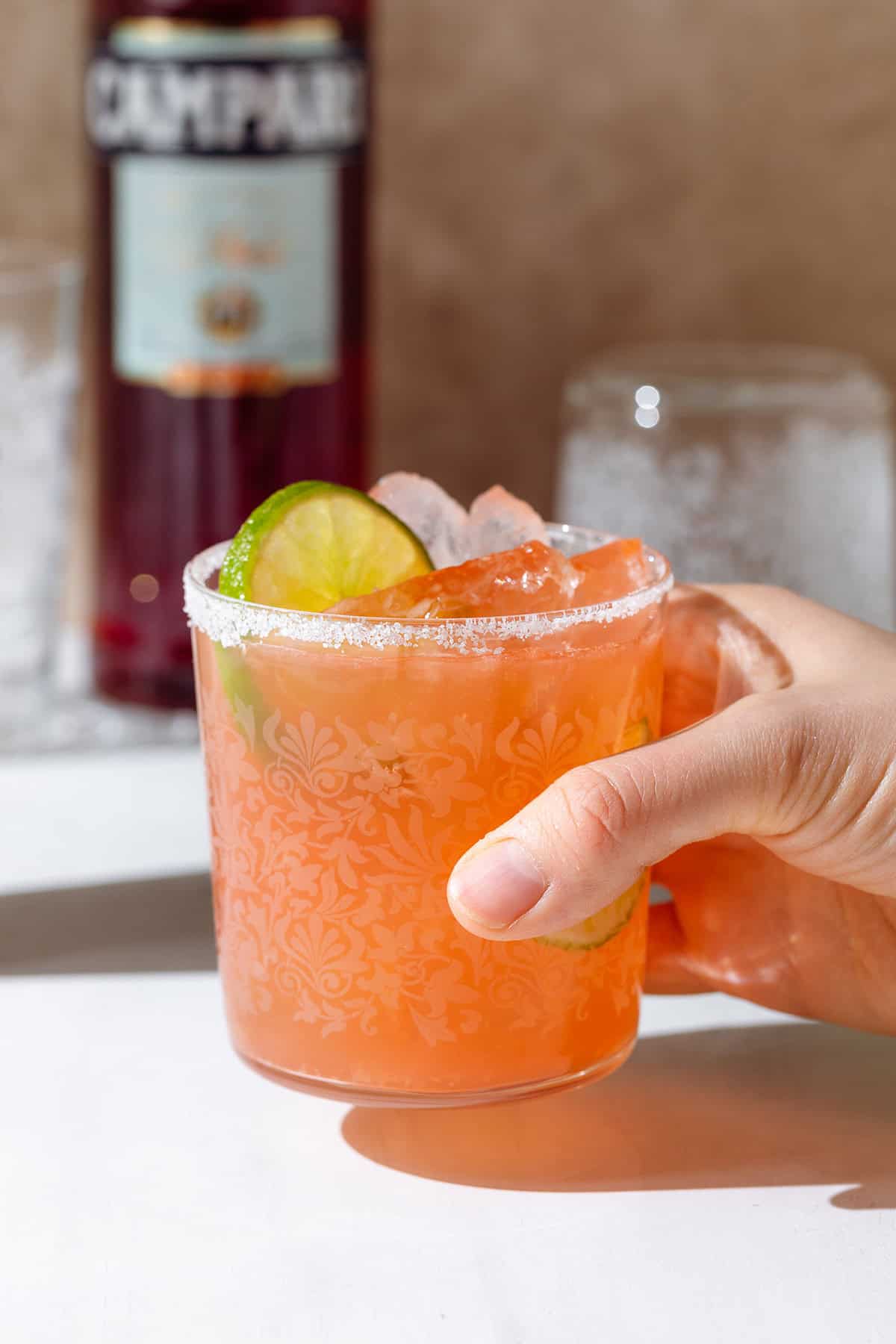 A hand holding an orange drink in a short glass filled with ice garnished with a lime slice and salt around the rim.