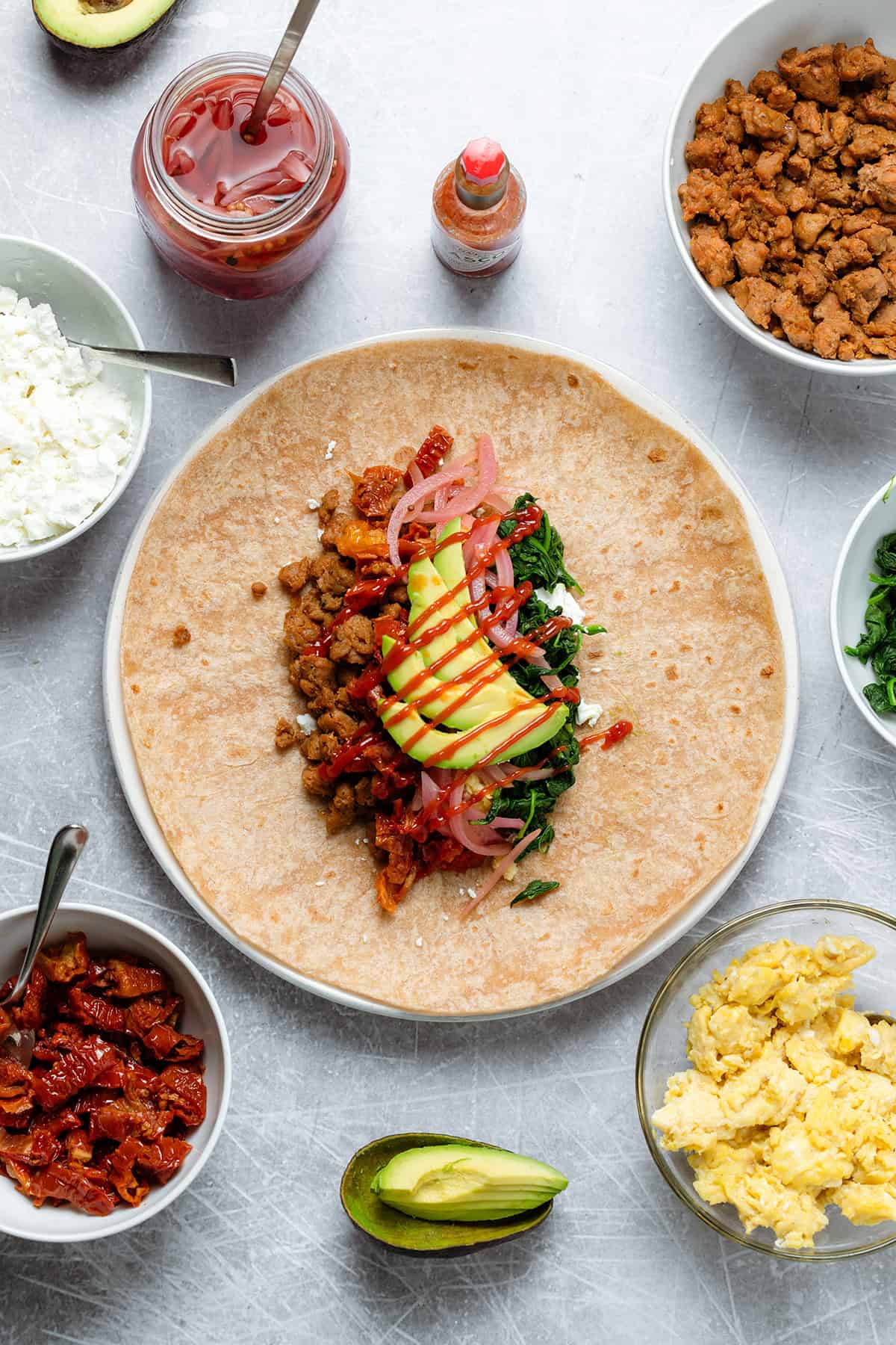 A tortilla wrap on a large plate with egg, sausage, avocado, and other ingredients on it ready to be wrapped.