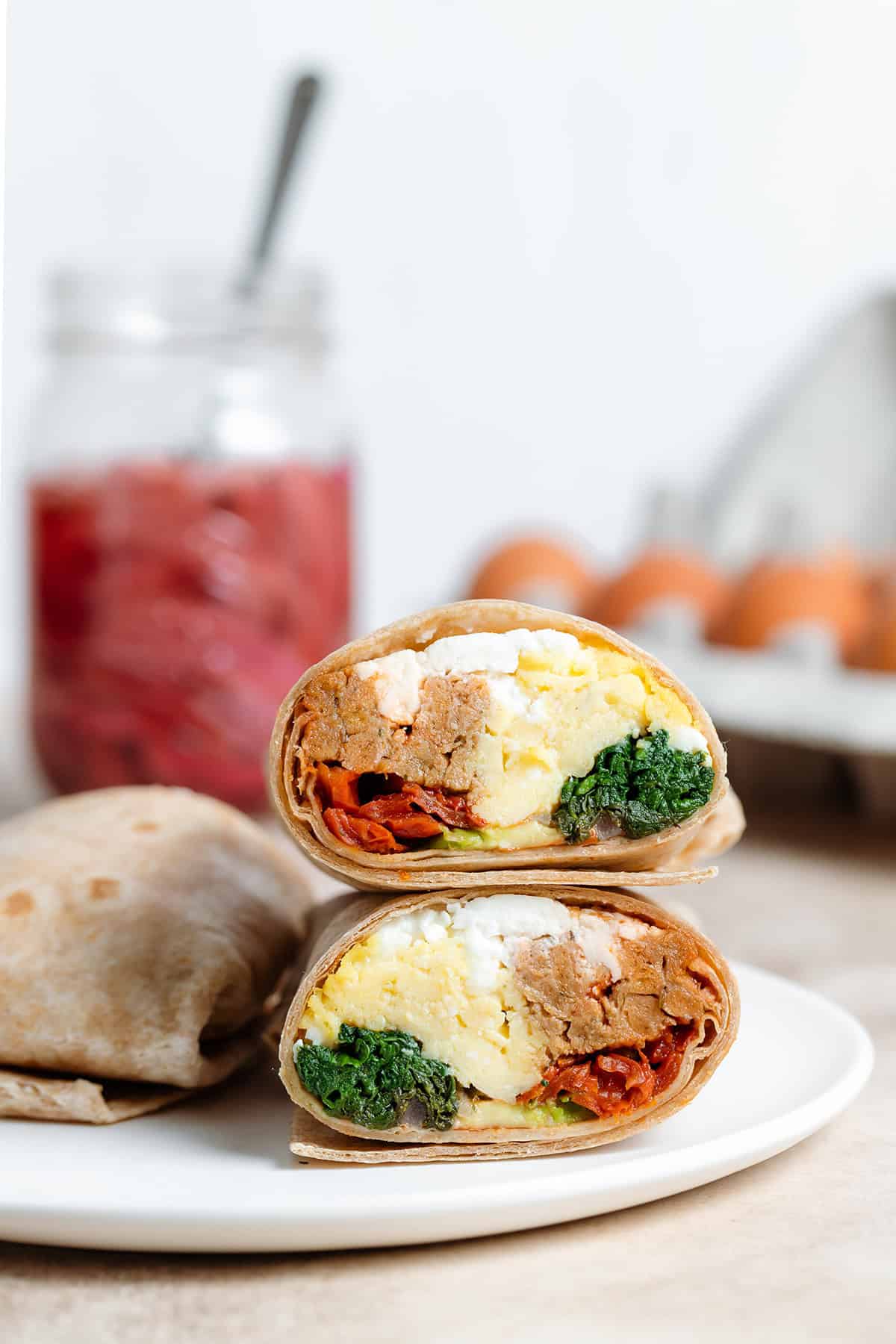 A breakfast burrito cut in half showing eggs, spinach, and sausage inside on a white plate with pickled onions and eggs in the background.