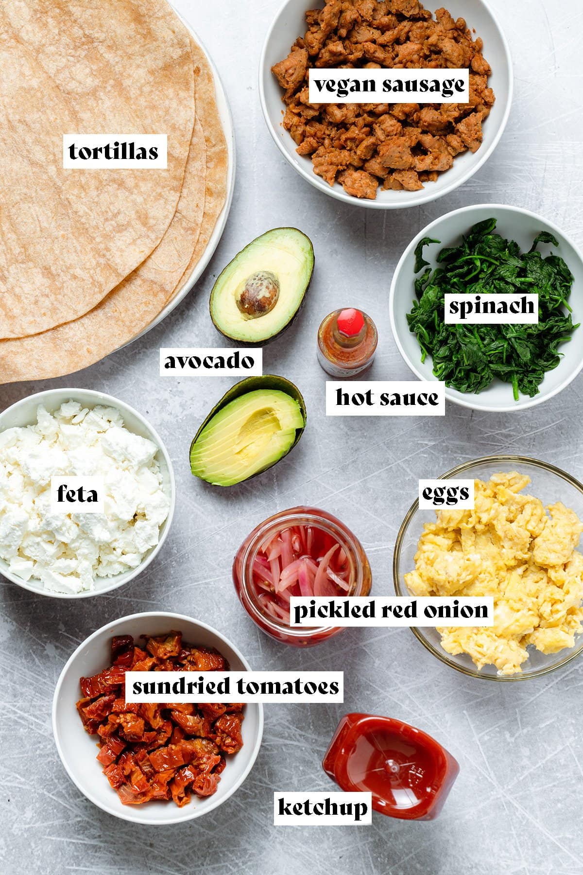 Ingredients like tortilla wraps, feta, spinach, and scrambled eggs laid out in bowls on metal background.
