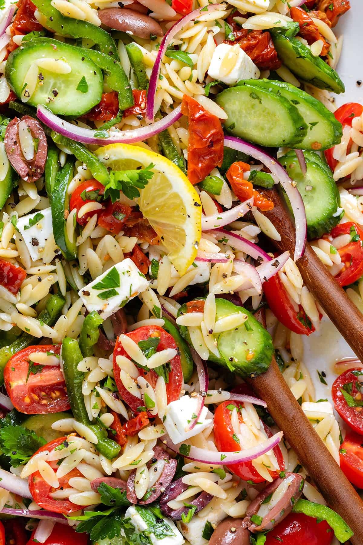 Greek orzo salad garnished with lemon slices on a large white platter with wooden serving spoons.