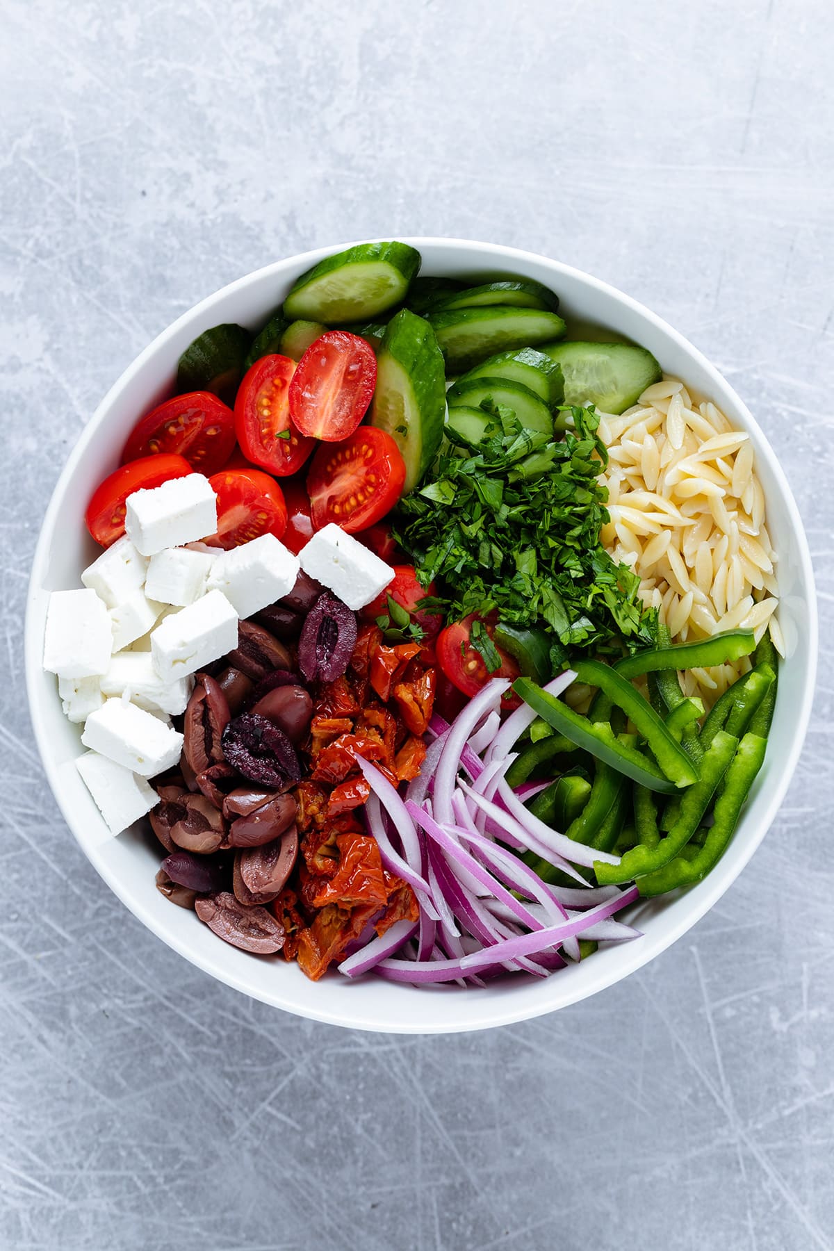 Ingredients for greek orzo salad like feta, olives, and vegetables separately laid out in a large bowl.