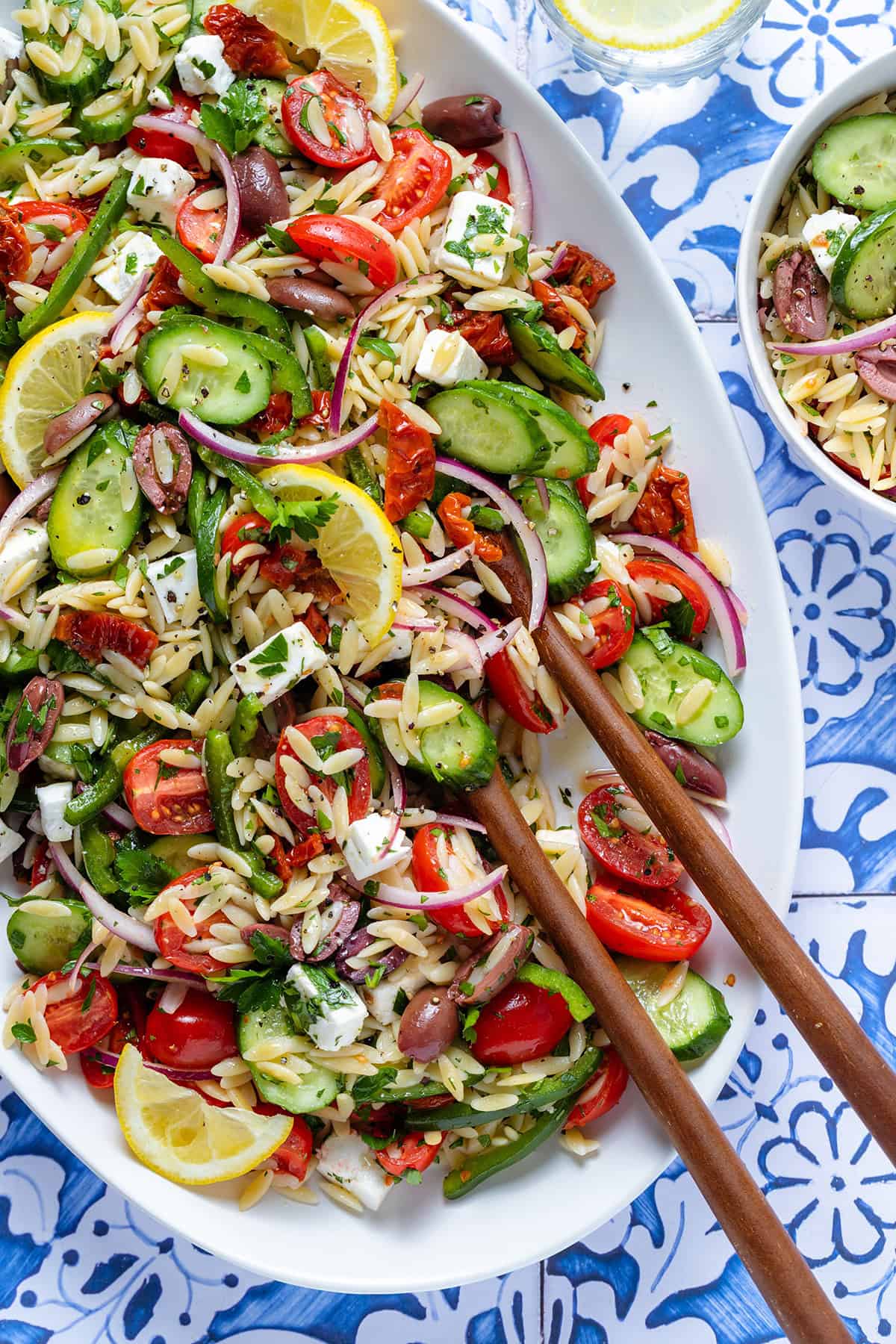 Greek orzo salad garnished with lemon slices on a large white platter with wooden serving spoons.