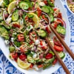 Greek orzo salad garnished with lemon slices on a large white platter with wooden serving spoons.