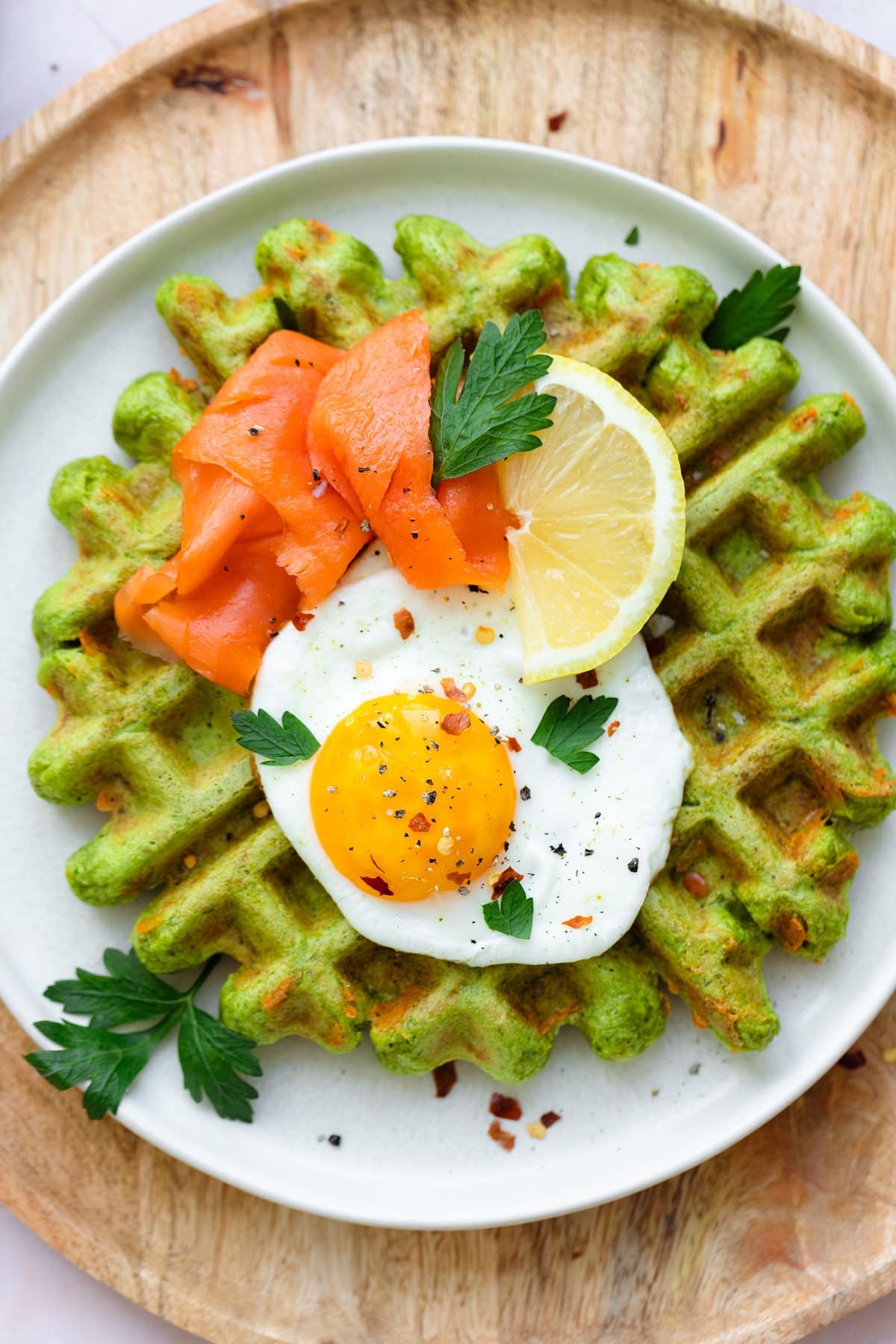 Green waffles on a white plate topped with a sunny side up egg, smoked salmon, fresh parsley, and a wedge of lemon.