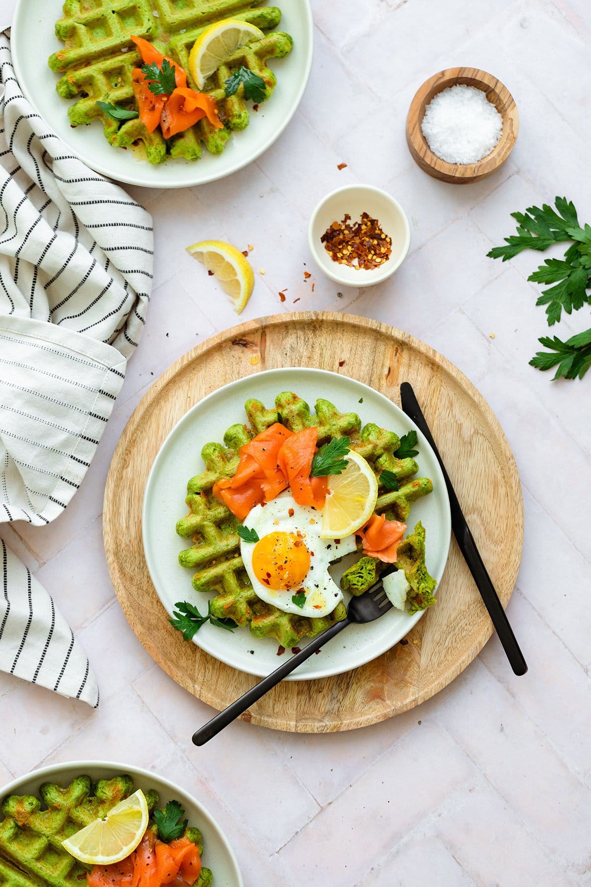 Green waffles on a white plate topped with a sunny side up egg that's been cut into, smoked salmon, fresh parsley, and a wedge of lemon.