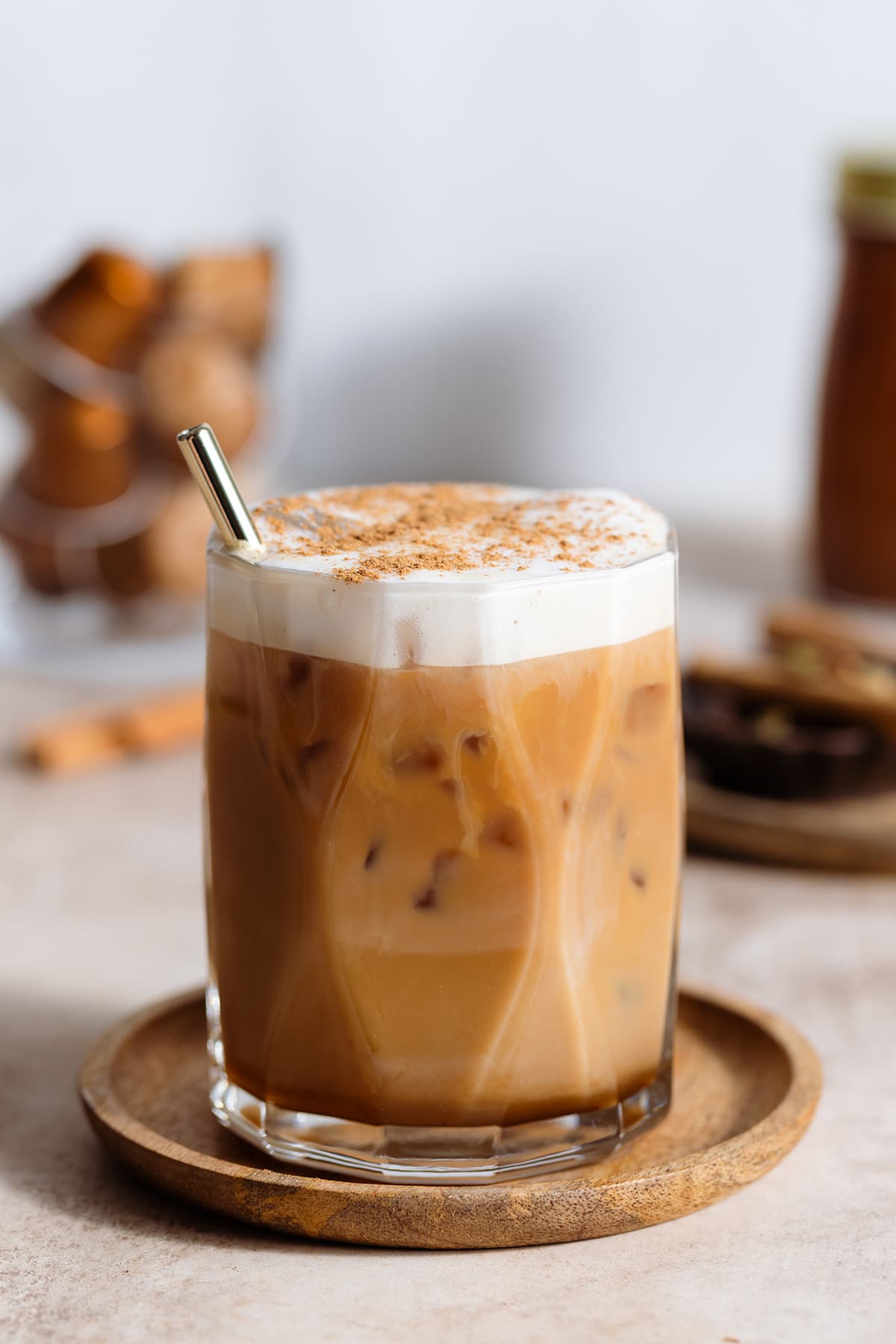 A glass with iced dirty chai latte and a gold straw on a wooden coaster and beige backdrop.