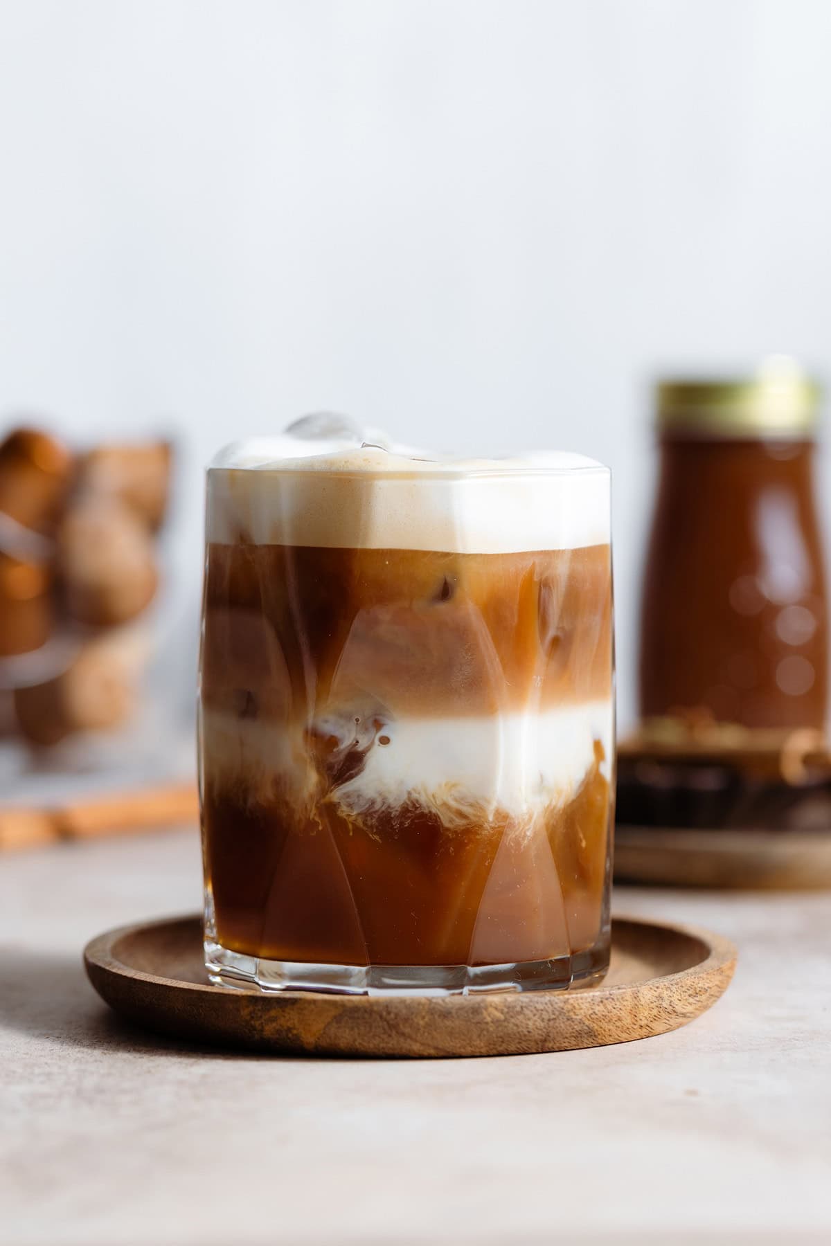 A glass with iced dirty chai latte with milk and chai layers separated on a wooden coaster and beige backdrop.