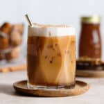 A glass with iced dirty chai latte and a gold straw on a wooden coaster and beige backdrop.