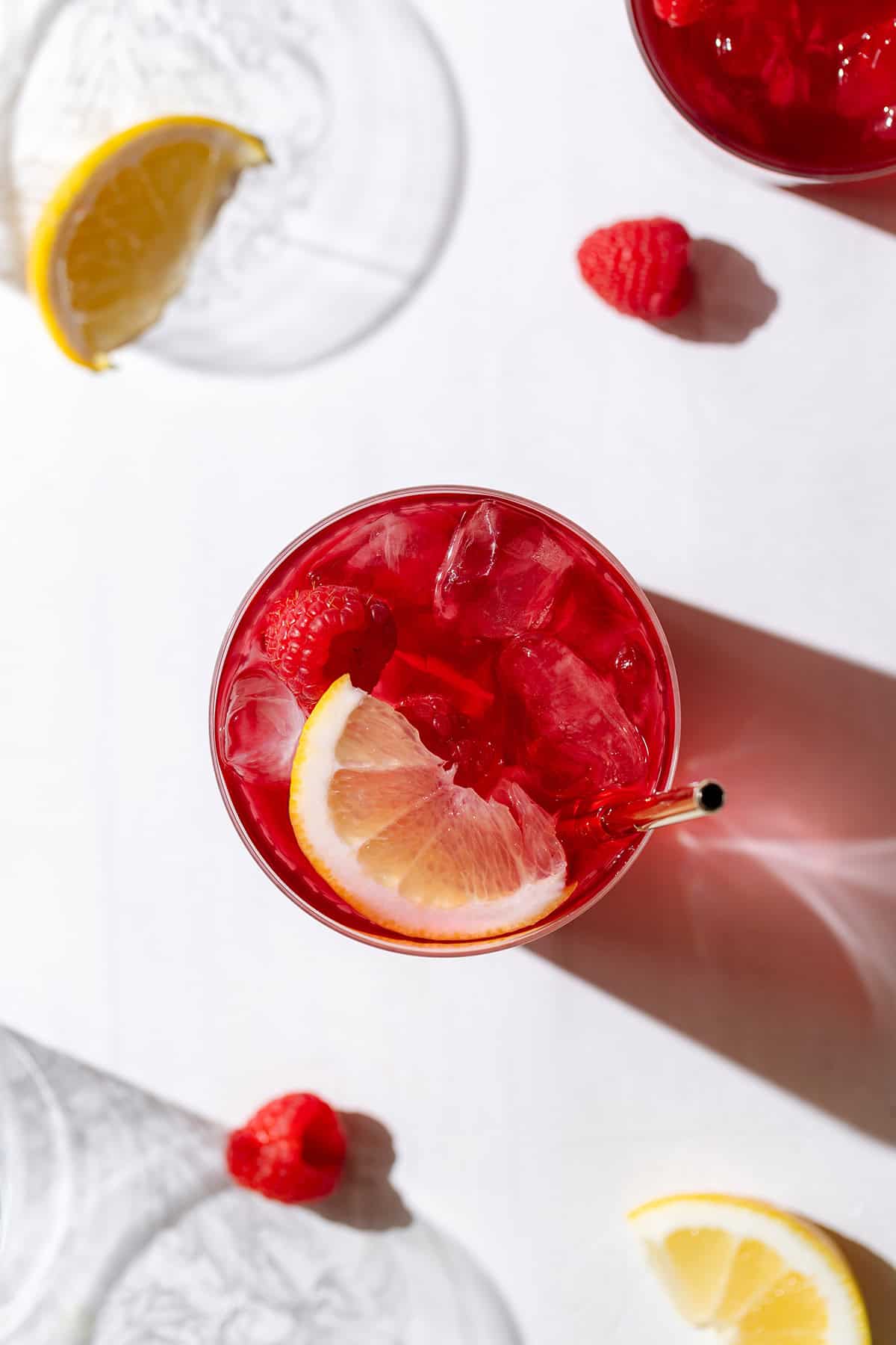 Iced red tea in a short glass with a gold metal straw garnished with a raspberry and a lemon wedge.