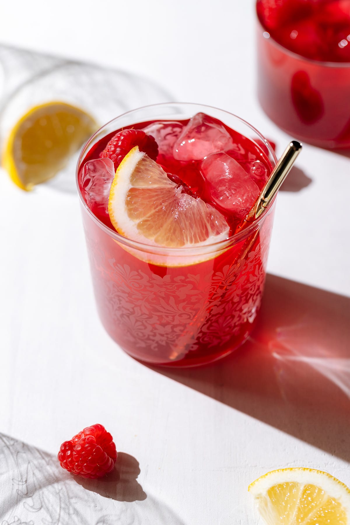 Iced red tea in a short glass with a gold metal straw garnished with a raspberry and a lemon wedge.