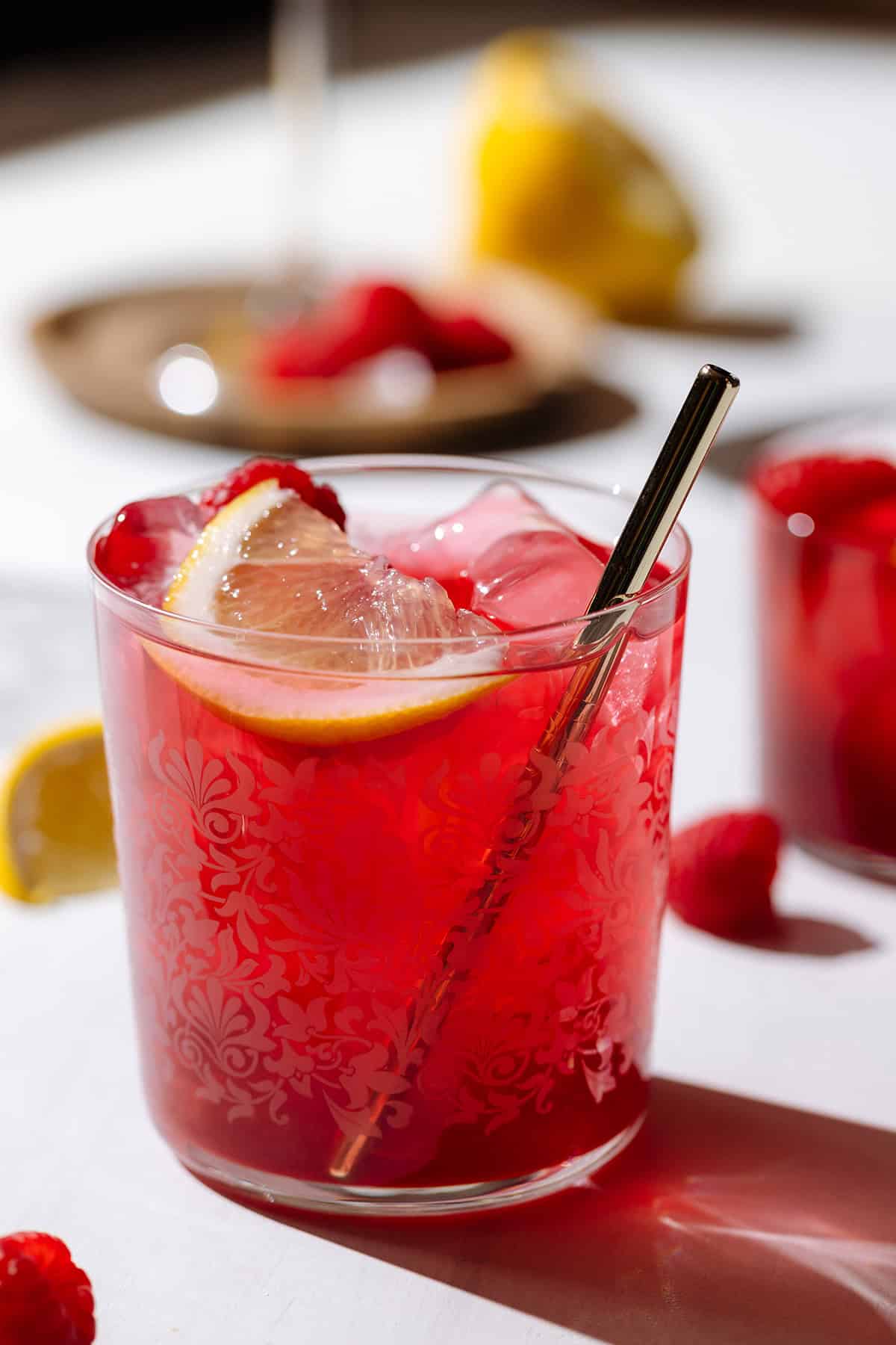 Iced red tea in a short glass with a gold metal straw garnished with a raspberry and a lemon wedge.