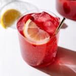 Iced red tea in a short glass with a gold metal straw garnished with a raspberry and a lemon wedge.