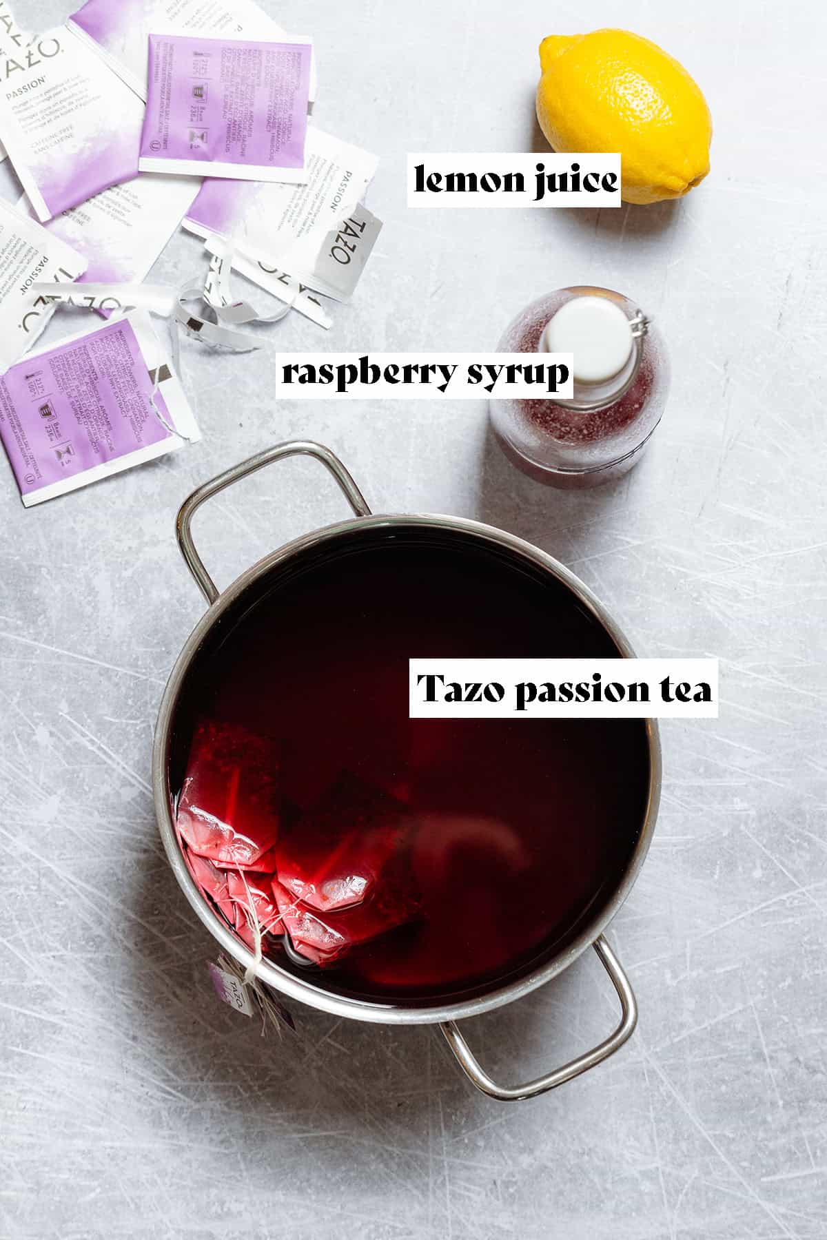 Red tea being steeped in a small pot with tea bags, lemon, and raspberry syrup around.