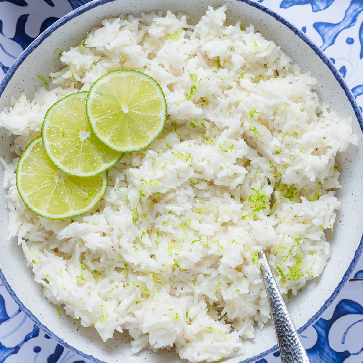 Coconut lime rice garnished with lime slices in a white bowl with a blue edge on a blue tile background.