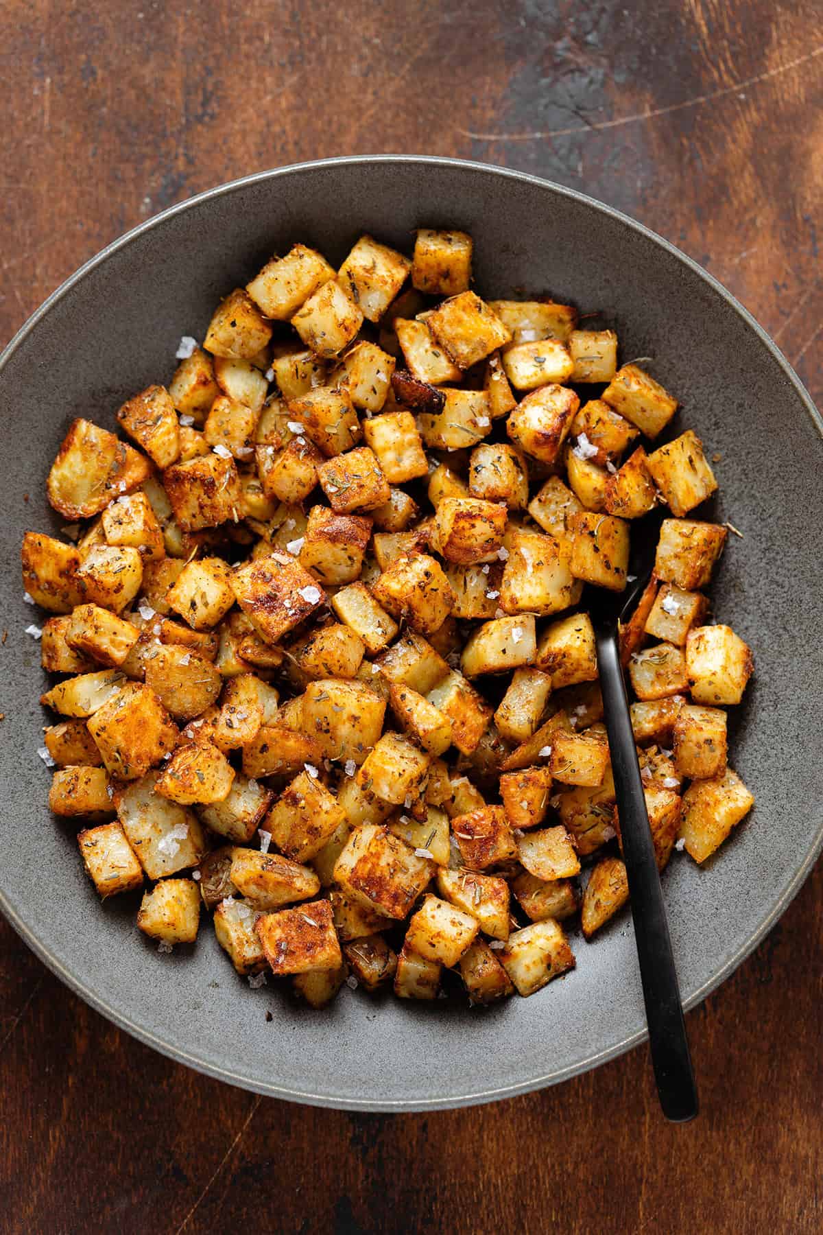 Roasted diced potatoes in a black bowl and a black serving spoon.