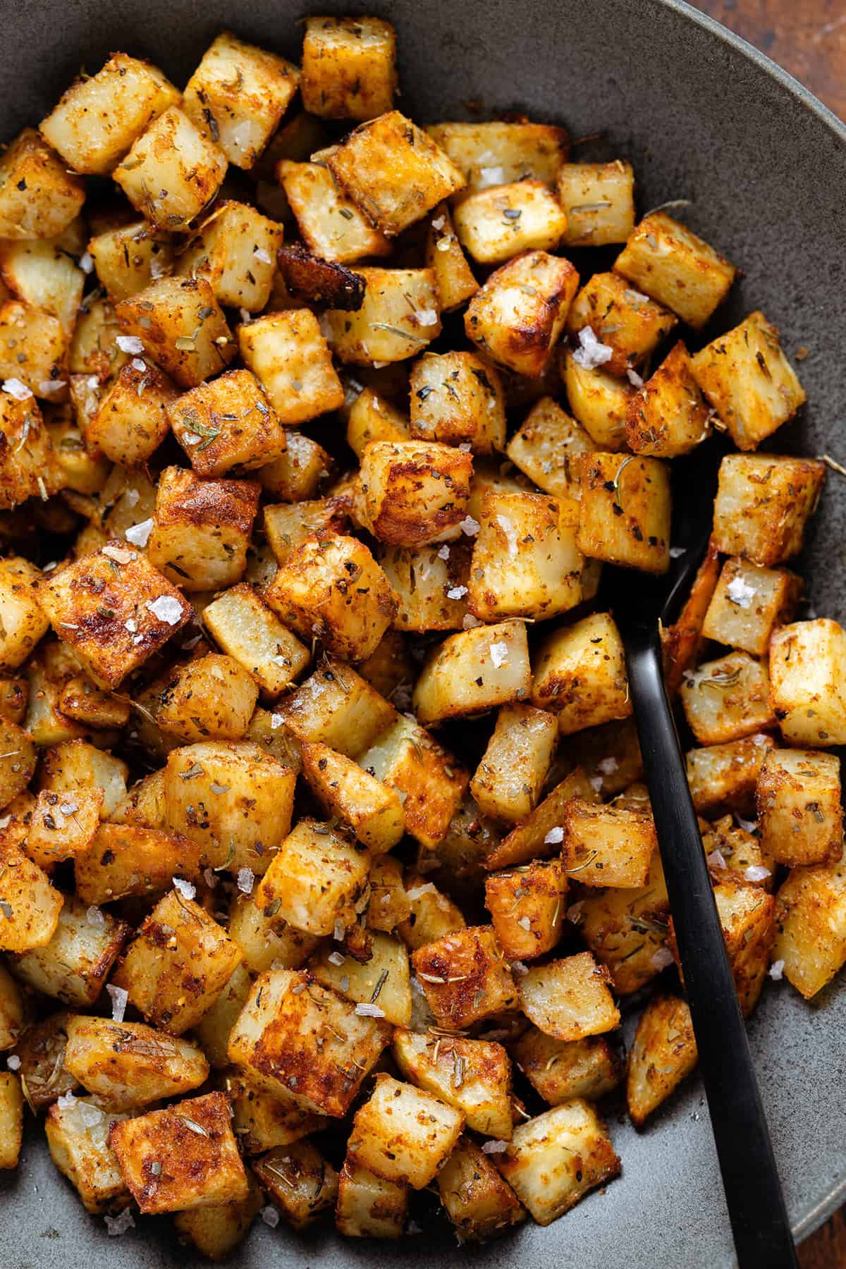 Roasted diced potatoes in a black bowl and a black serving spoon.