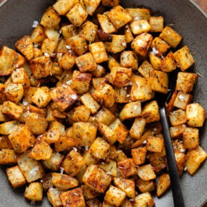 Roasted diced potatoes in a black bowl and a black serving spoon.