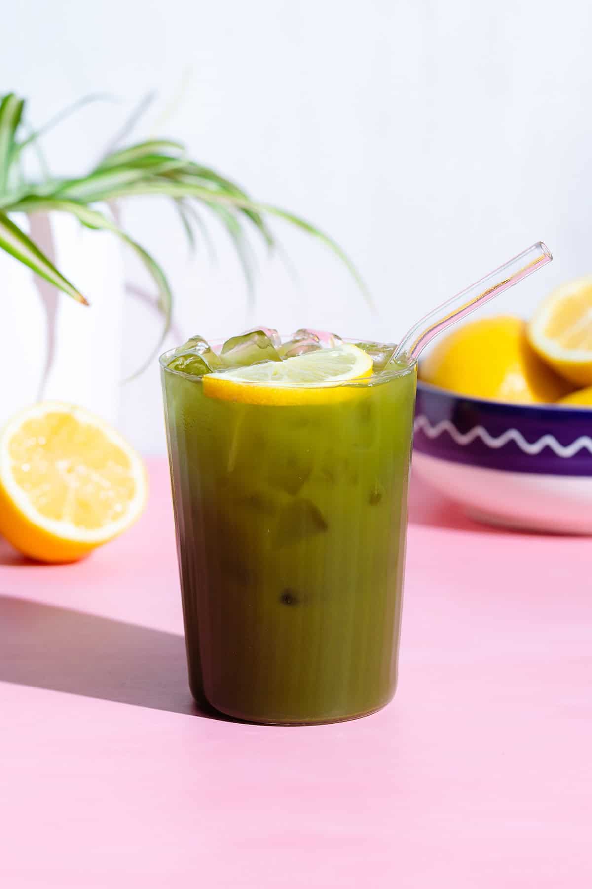 Green Matcha lemonade with a lemon slice in a tall glass with a glass straw on a pink background.