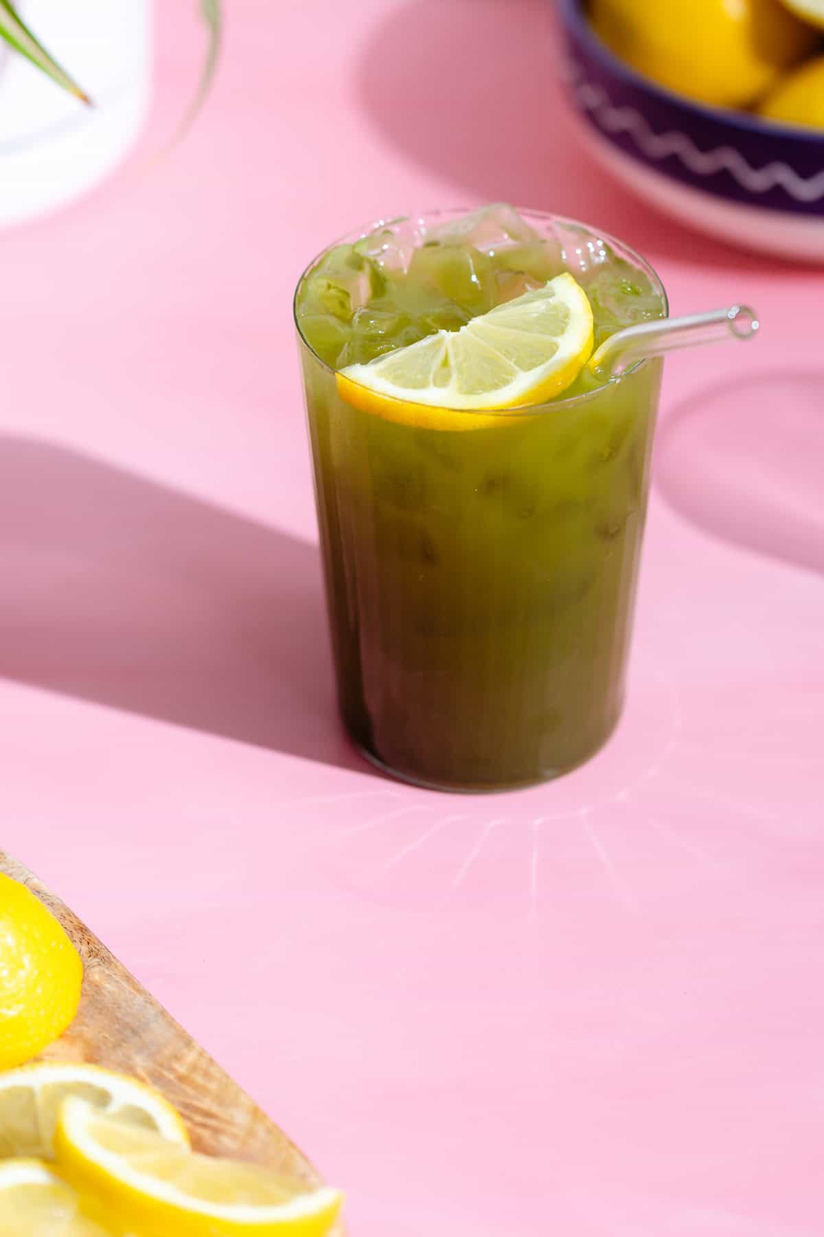 Matcha lemonade with a lemon slice in a tall glass with a glass straw on a pink background.