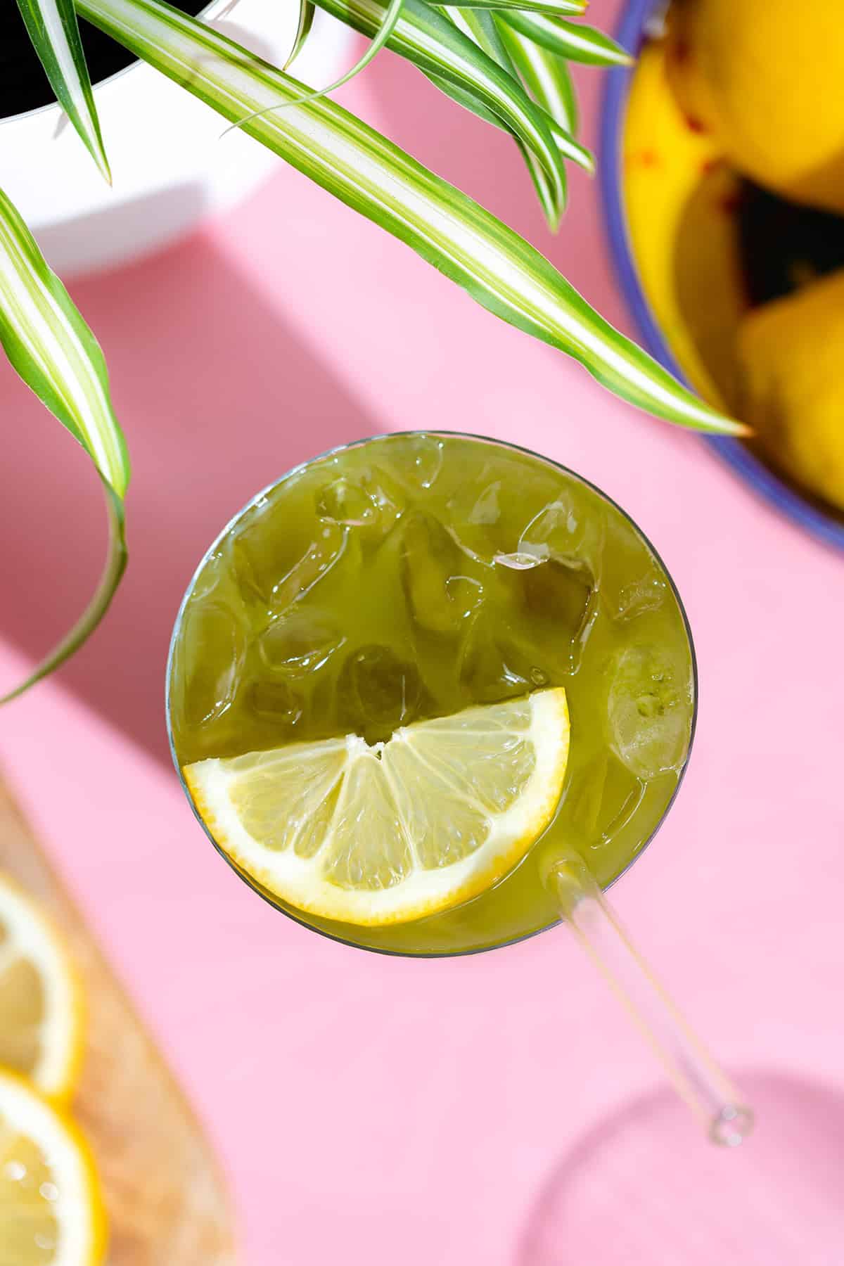 Matcha lemonade with a lemon slice in a tall glass with a glass straw on a pink background.