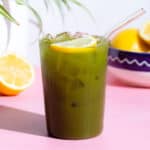 Matcha lemonade with a lemon slice in a tall glass with a glass straw on a pink background.