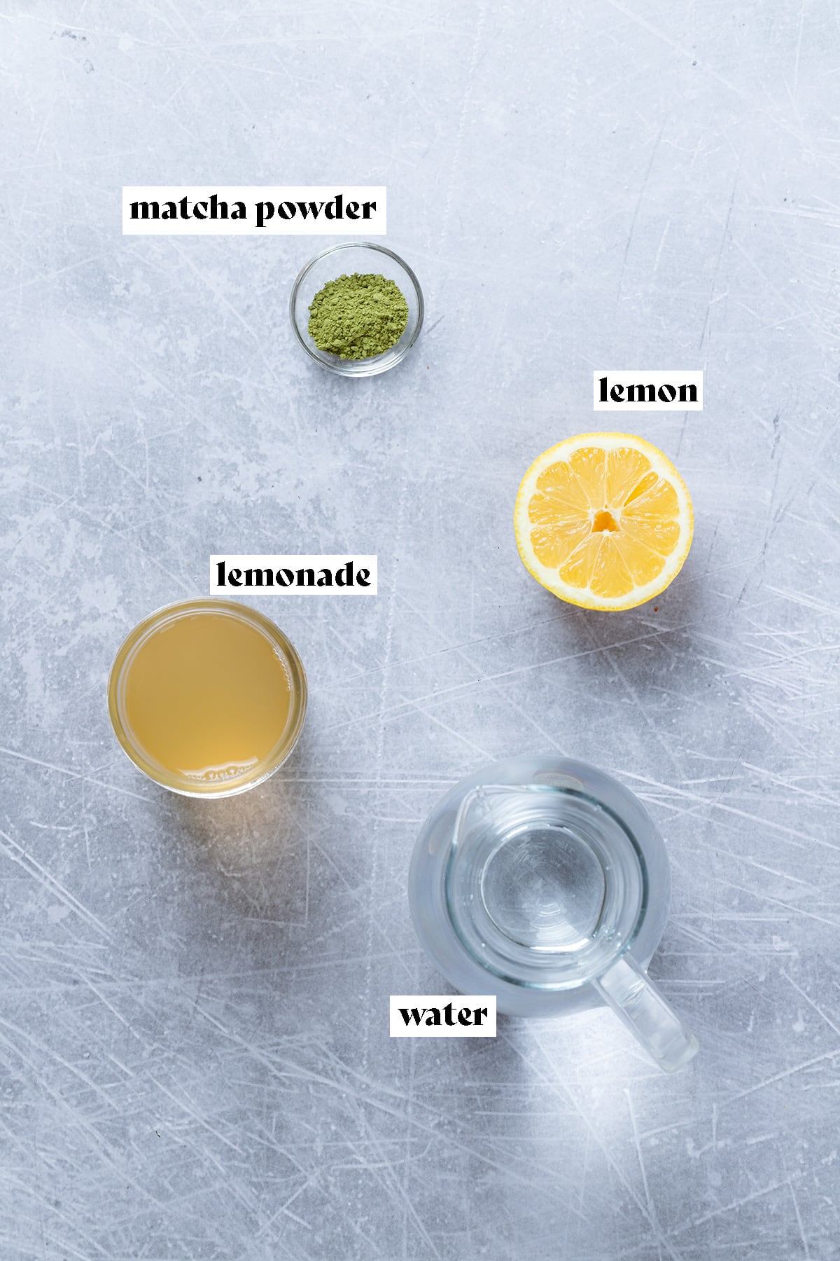 Matcha powder, lemonade, water, and a lemon laid out on a metal background.