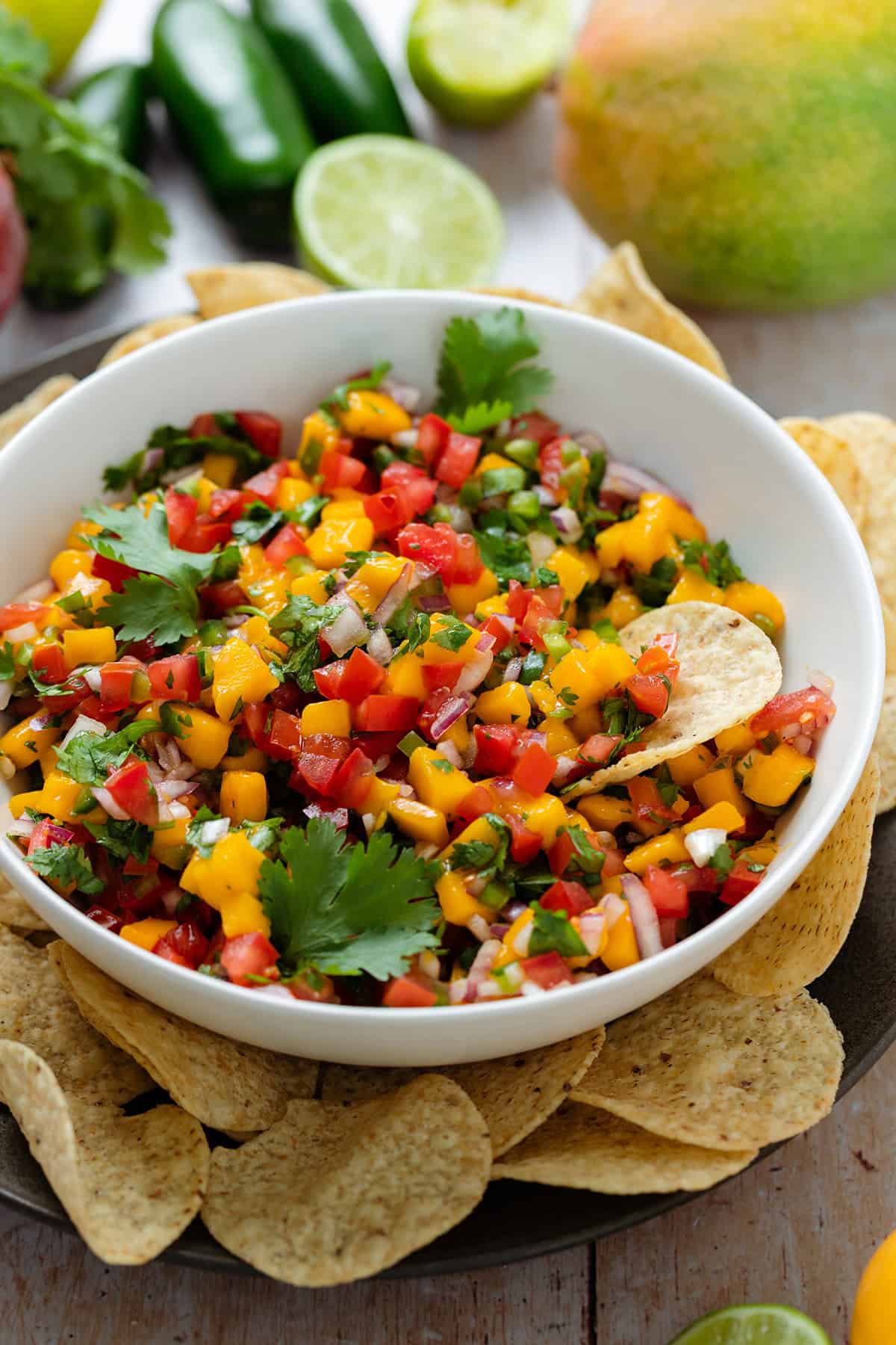 Mango pico de gallo in a low white bowl on a black plate with round tortilla chips around the bowl.