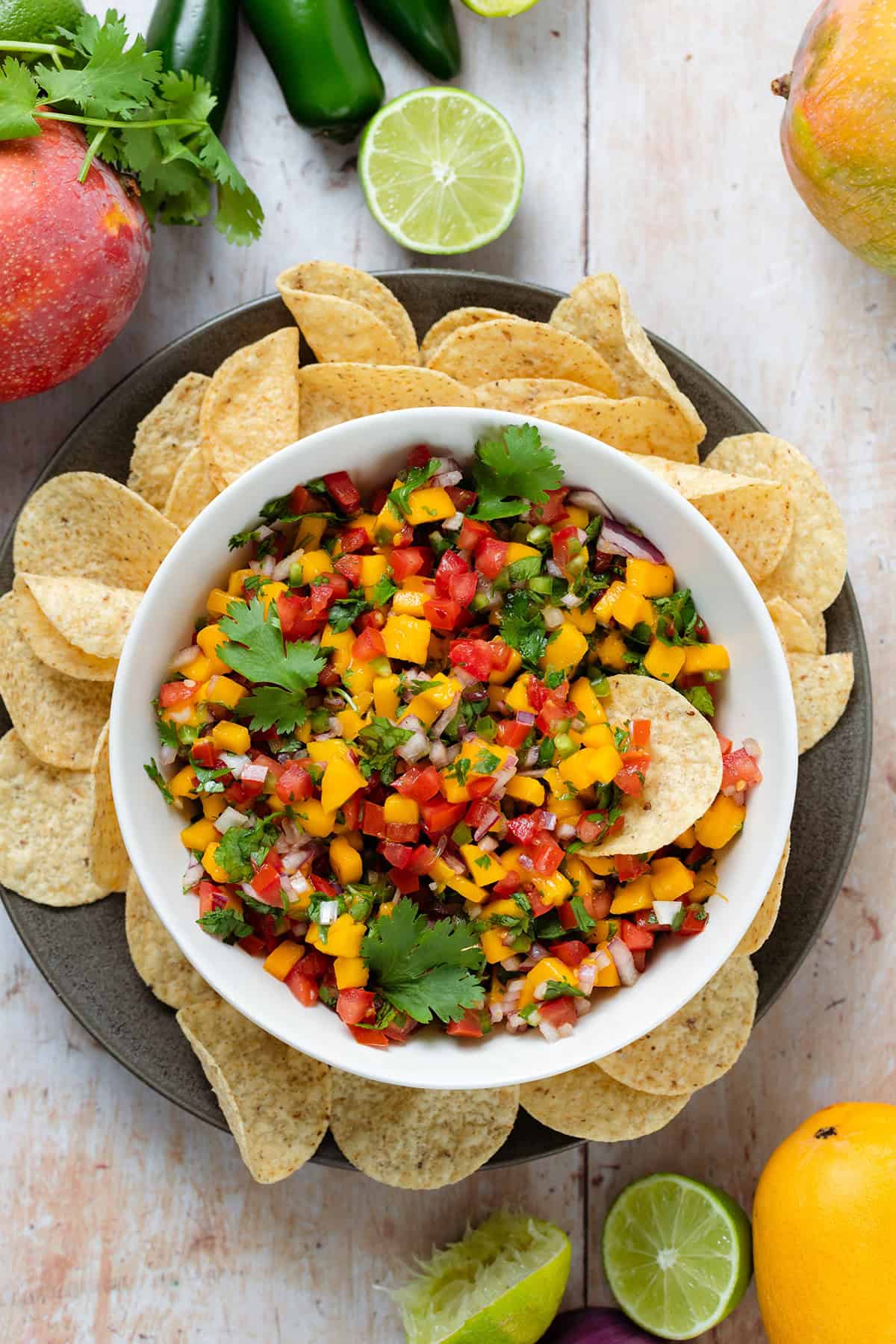 Mango pico de gallo in a low white bowl on a black plate with round tortilla chips around the bowl.