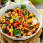 Mango pico de gallo in a low white bowl on a black plate with round tortilla chips around the bowl.