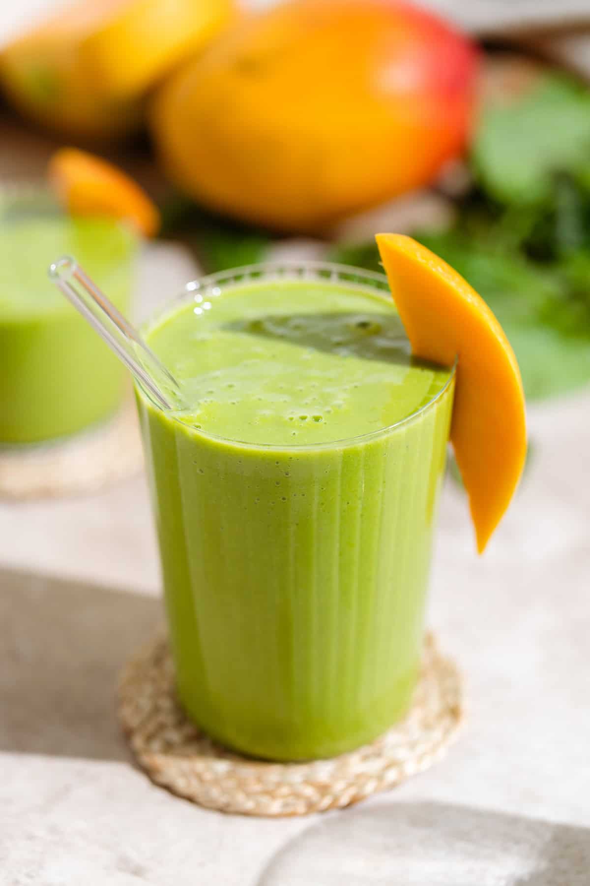 Green smoothie in a tall glass garnished with a slice of mango on a woven coaster with mangos and spinach in the background.