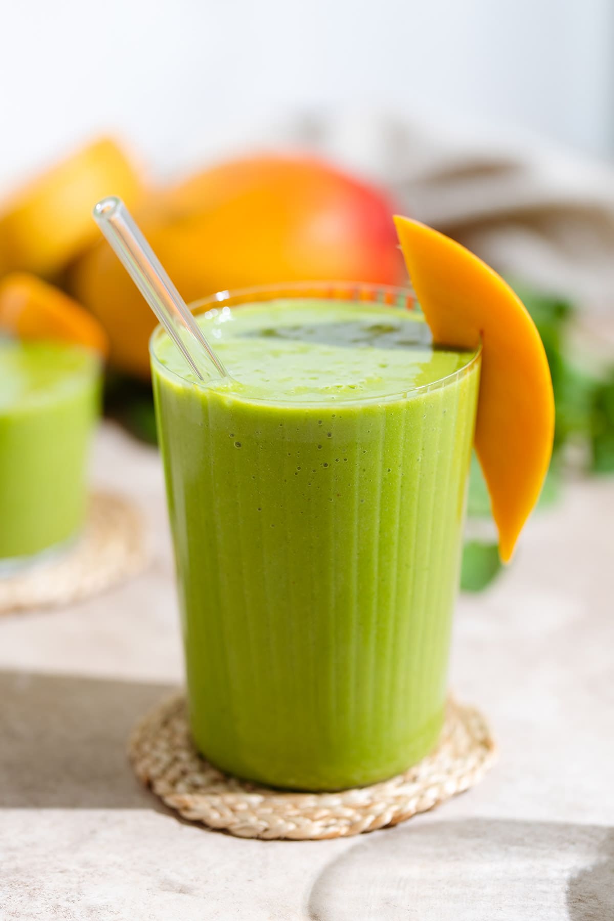 Green smoothie in a tall glass garnished with a slice of mango on a woven coaster with mangos and spinach in the background.