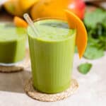 Green smoothie in a tall thin wall glass garnished with a slice of mango on a woven coaster with mangos and spinach in the background.